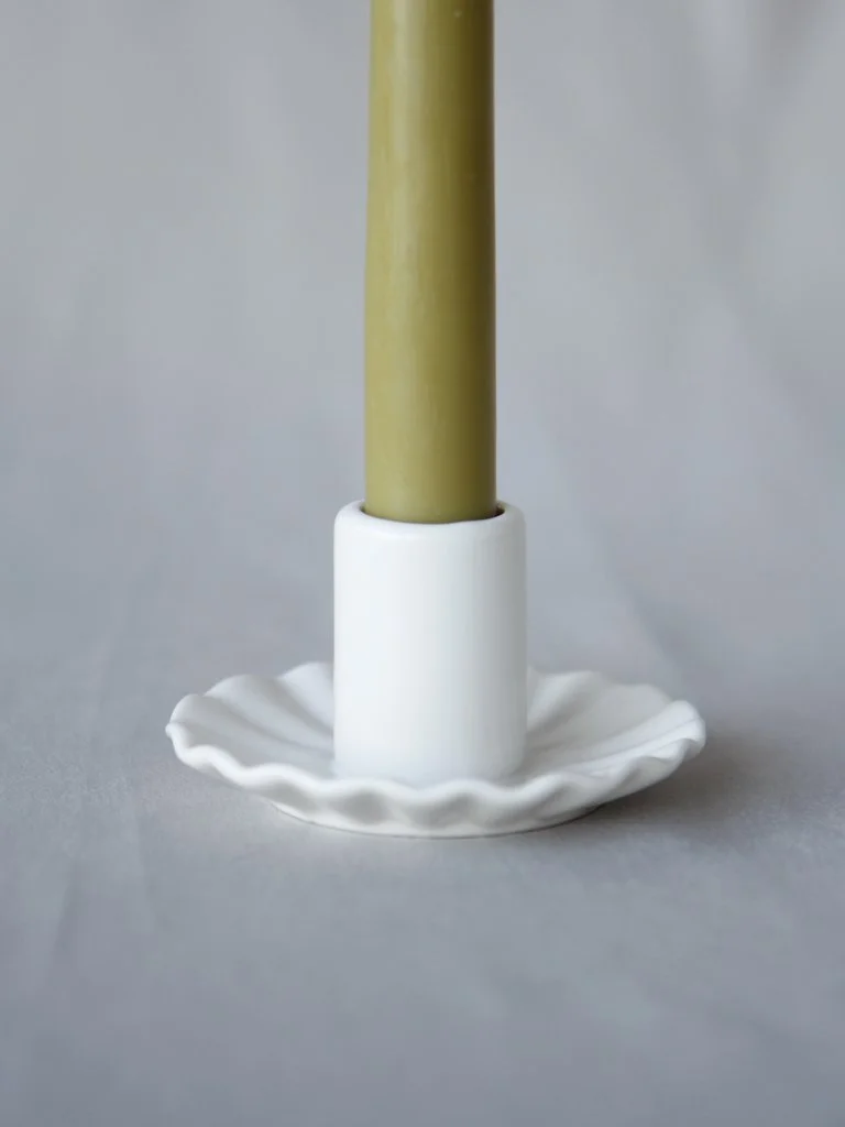 A green candle in a white candle holder on a white dish against a neutral background.