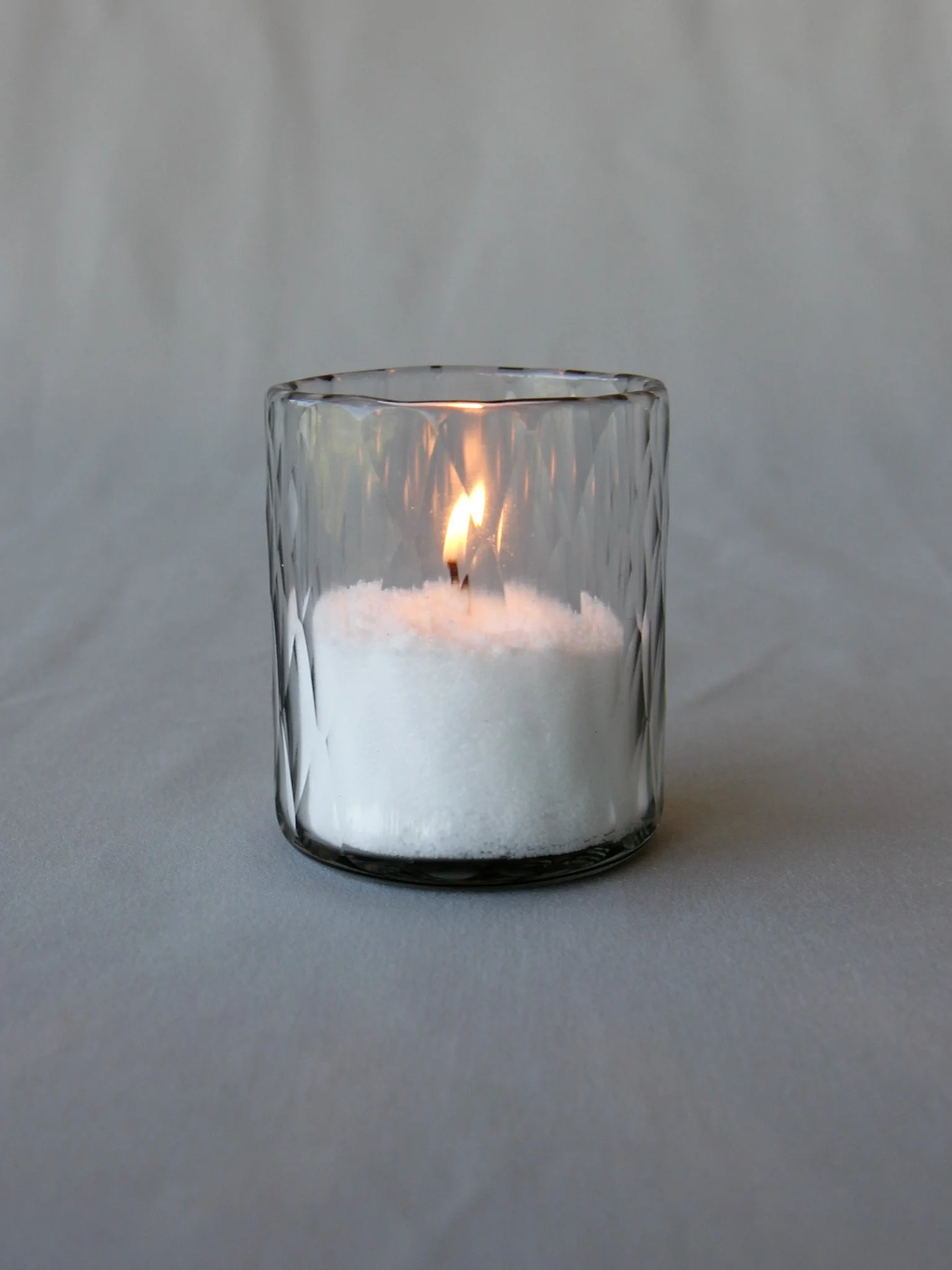 A lit white candle in a textured clear glass holder on a gray surface.