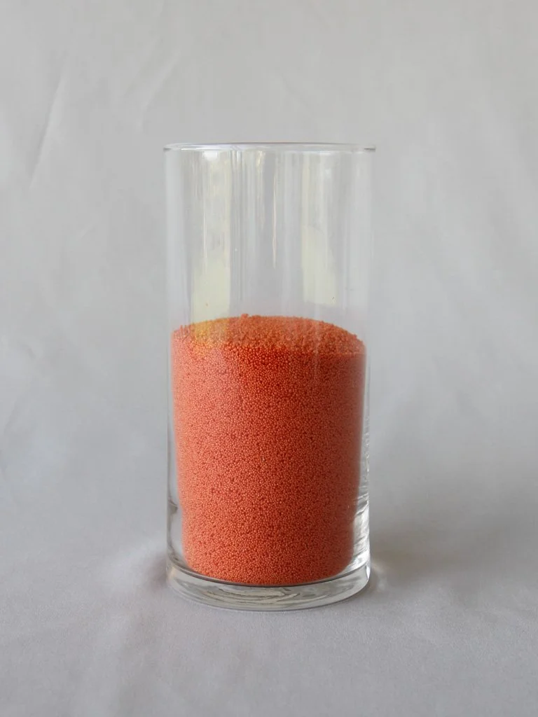 A clear glass filled with orange-colored small spherical beads against a plain background.