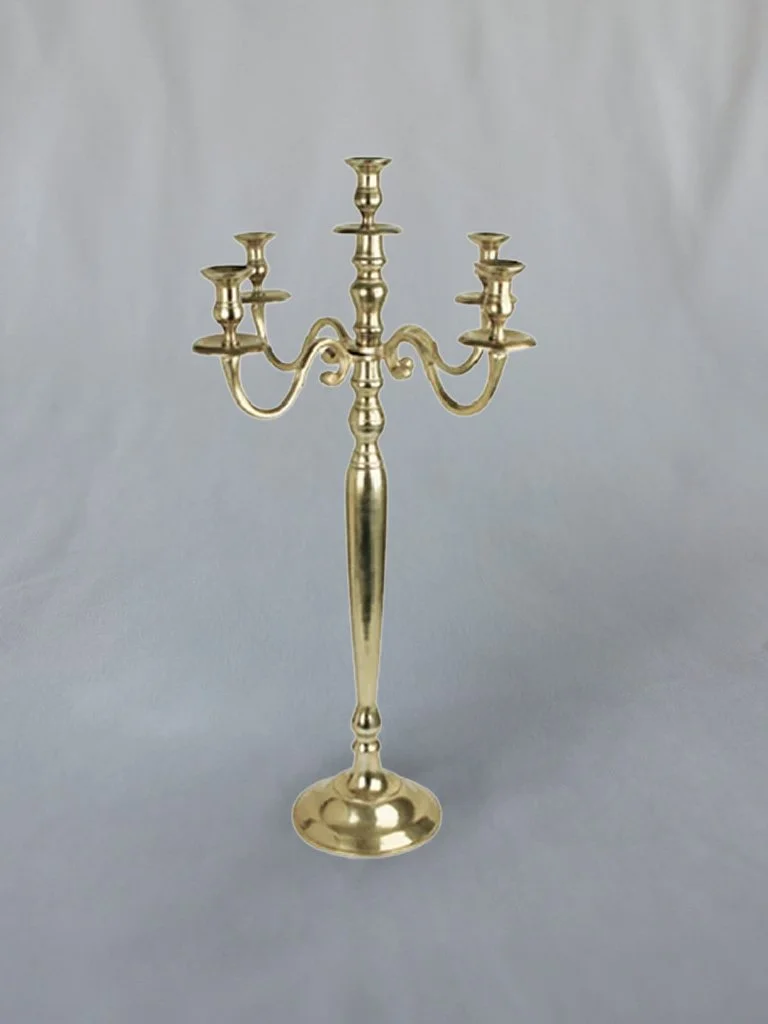 A gold-colored metal candelabrum with five candle holders, central support, and a rounded base, standing on a light gray surface against a plain light background.