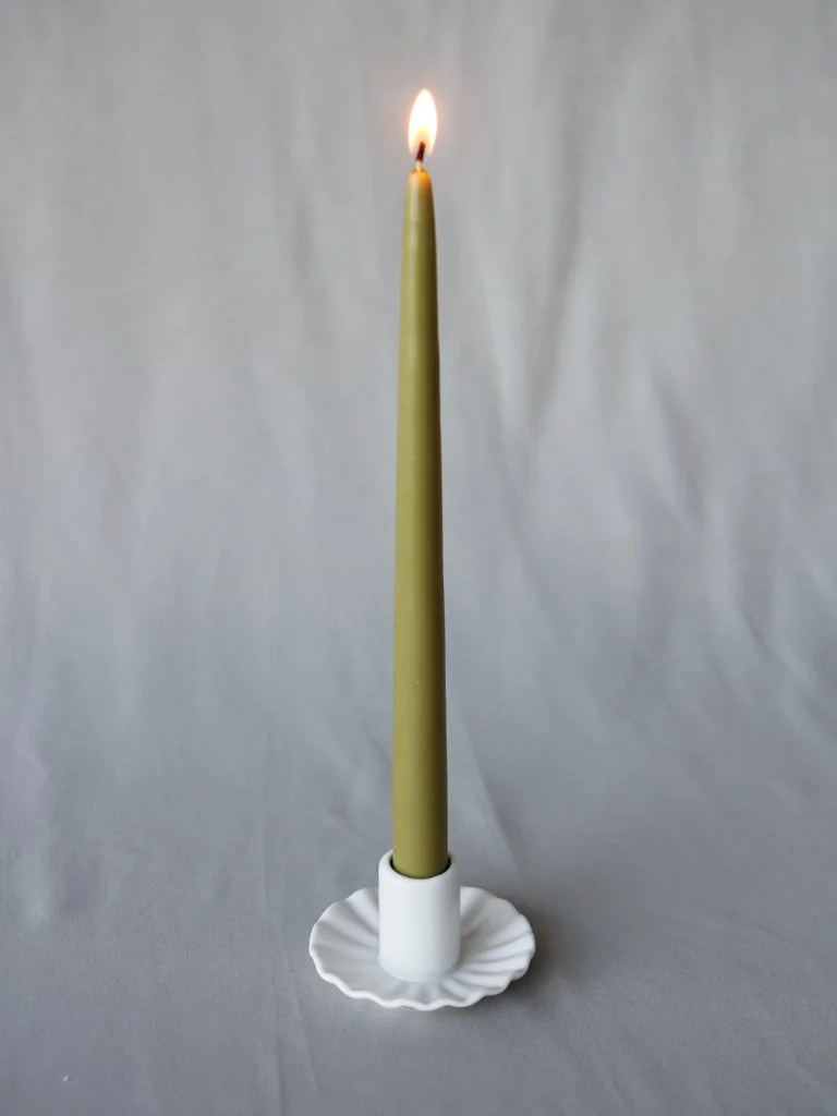 A tall yellow candle with a small flame in a white candle holder on a white textured surface.