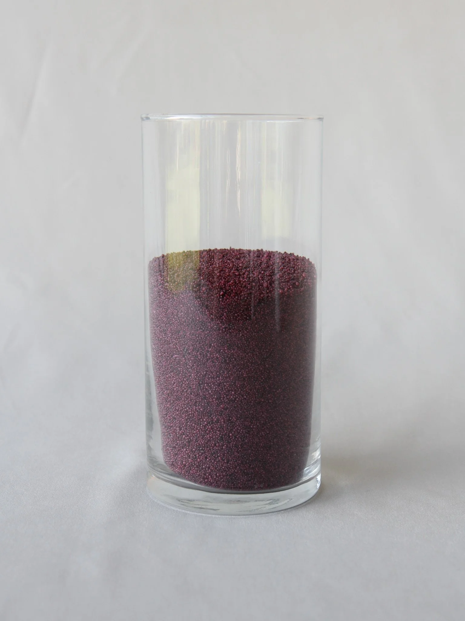 A tall, clear glass filled with purple tiny spherical beads, with some green beads on the top left.