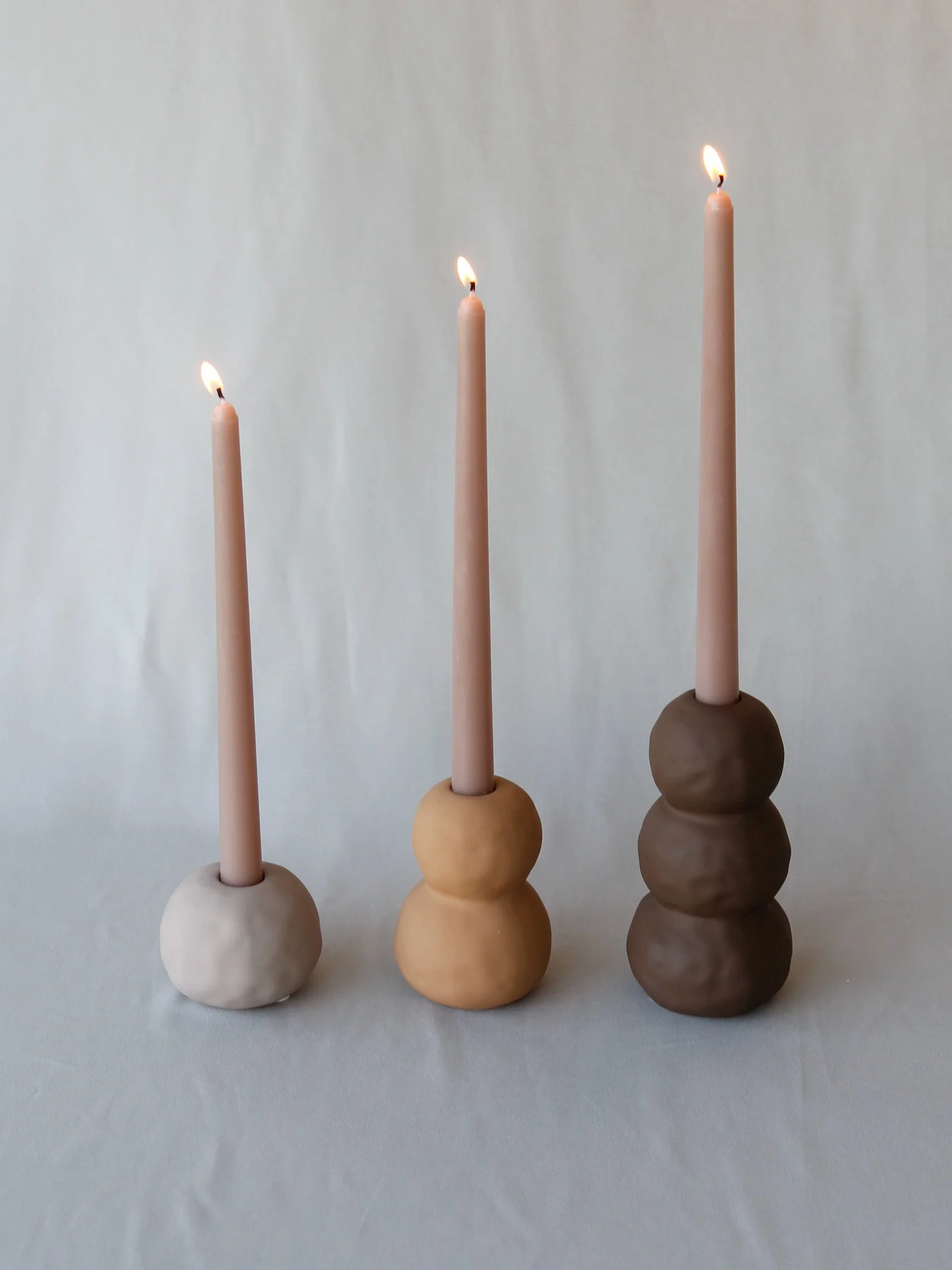 Three candles with pink bases of increasing height, lit and placed on round, stacked bases of different shades of beige and brown, on a white surface with a white background.