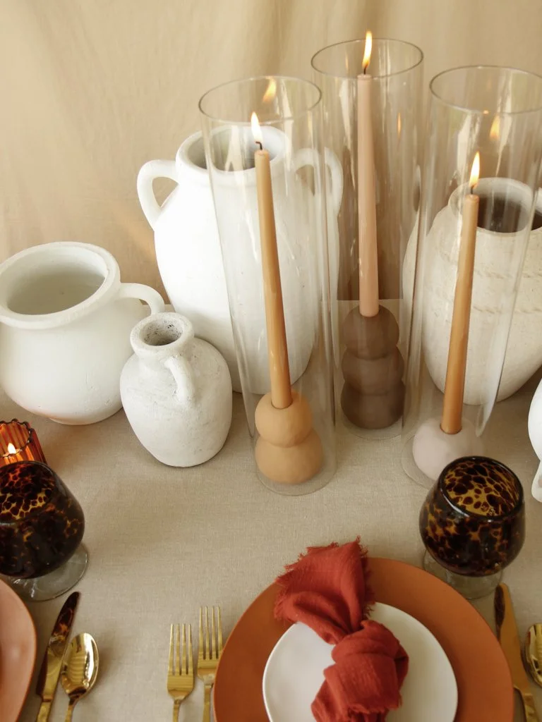 Decorative table setting with white ceramic vases, tall glass candle holders with lit candles, black and brown leopard print glasses, gold utensils, a white plate with a rust-colored napkin, and a beige tablecloth.