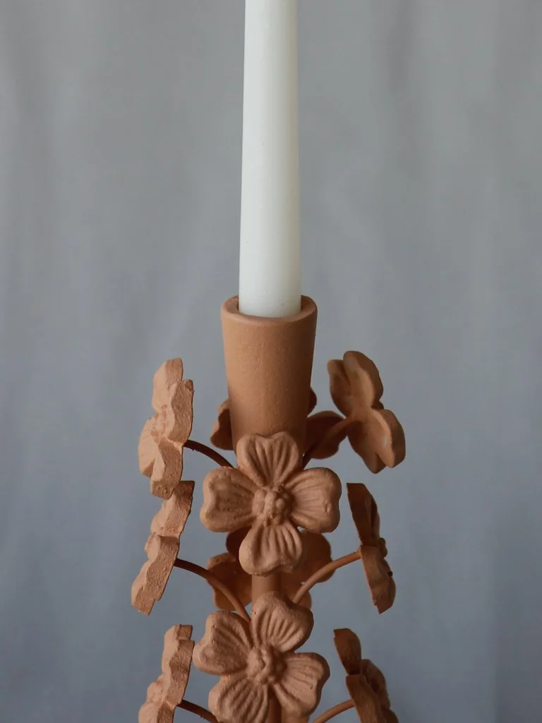A white candle is held in a light brown candle holder decorated with 3D flower and leaf designs, set against a plain light gray background.
