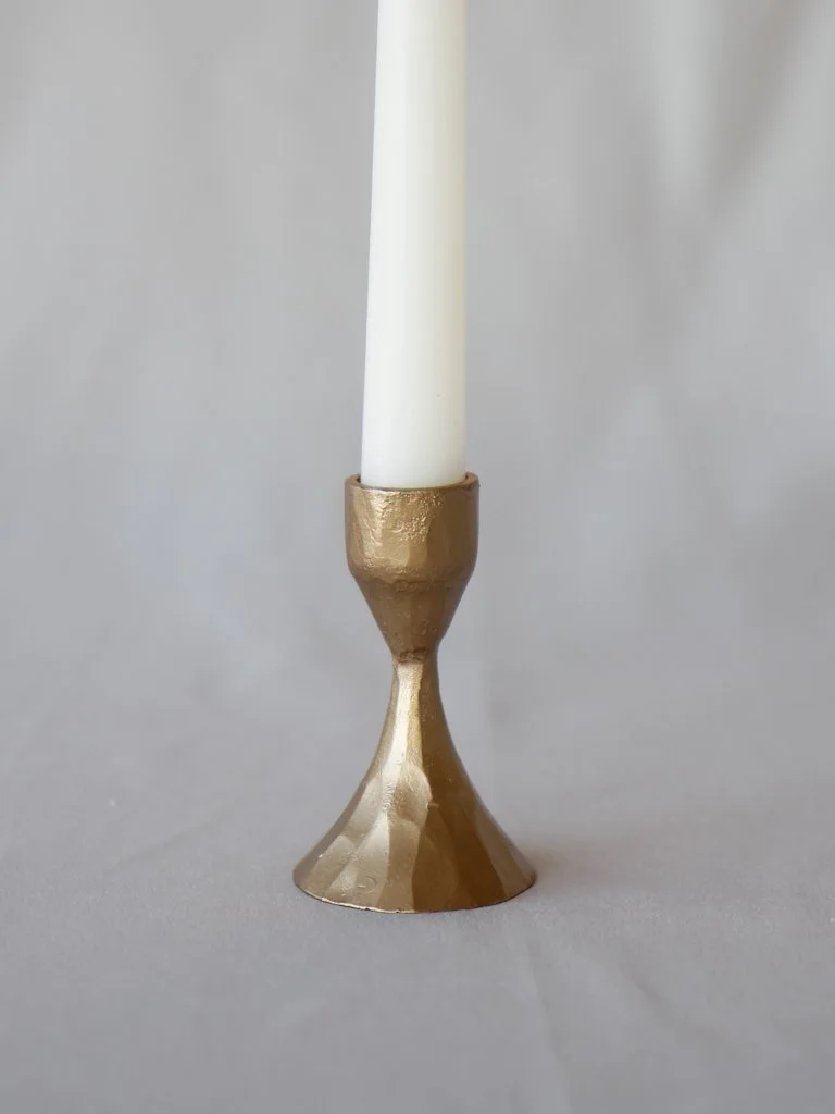 A candle holder with a metallic gold finish holding a white taper candle against a plain gray background.