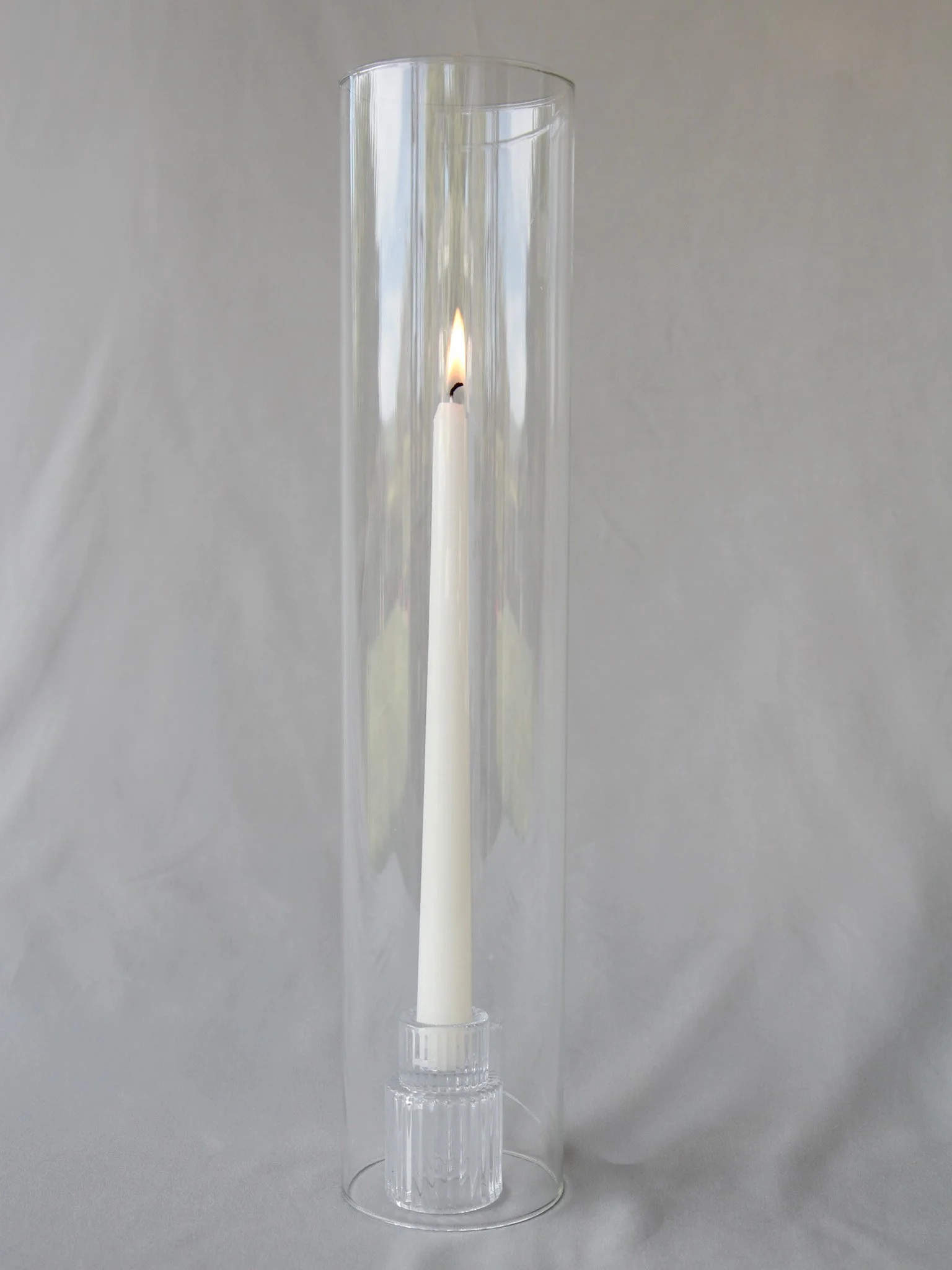 A tall, clear glass cylinder vase with a candle inside, a lit white candle with a small flame, positioned against a plain gray background.