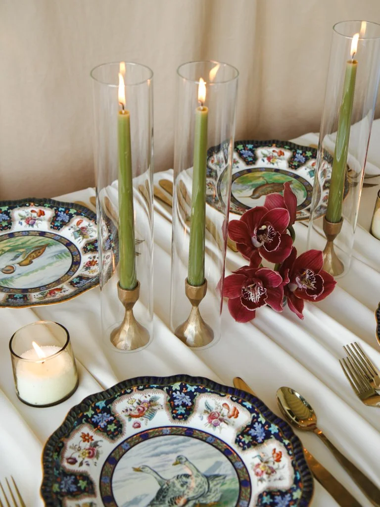 A beautifully set table with three tall glass candle holders with lit candles, a small lit candle in a glass holder, decorative plates with bird and floral designs, a bunch of red orchids, and gold utensils on a white tablecloth.
