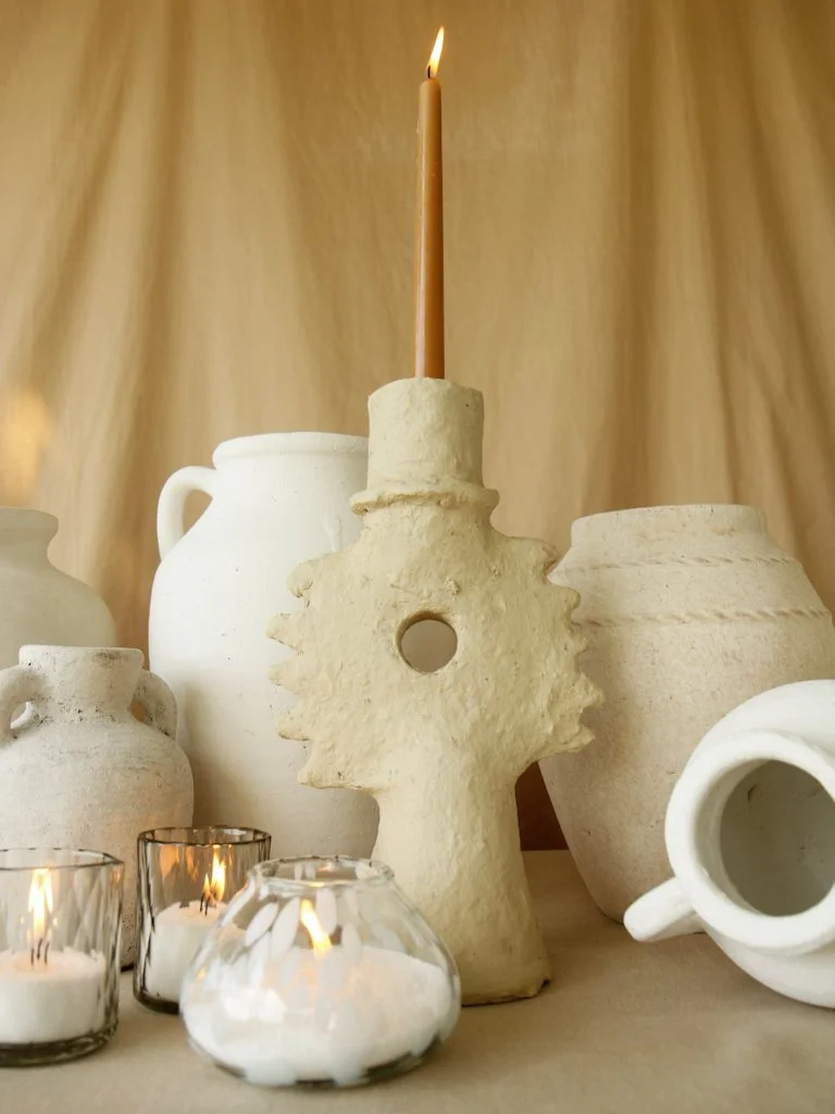 A lit candle on a beige candle holder surrounded by white and off-white ceramic vases and candle holders on a table with a beige background.