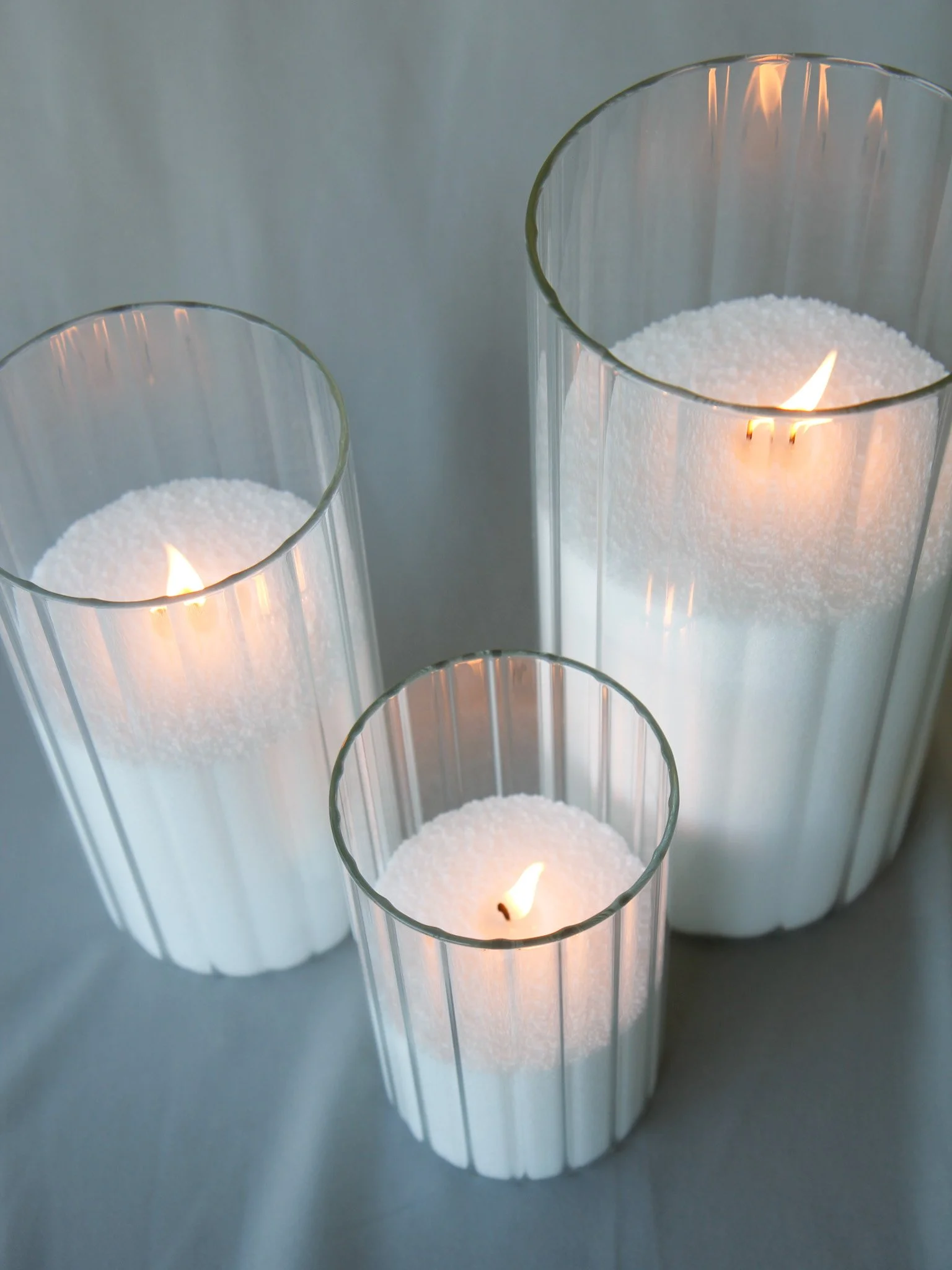 Three cylindrical glass candle holders with white candles inside, lit and burning softly.