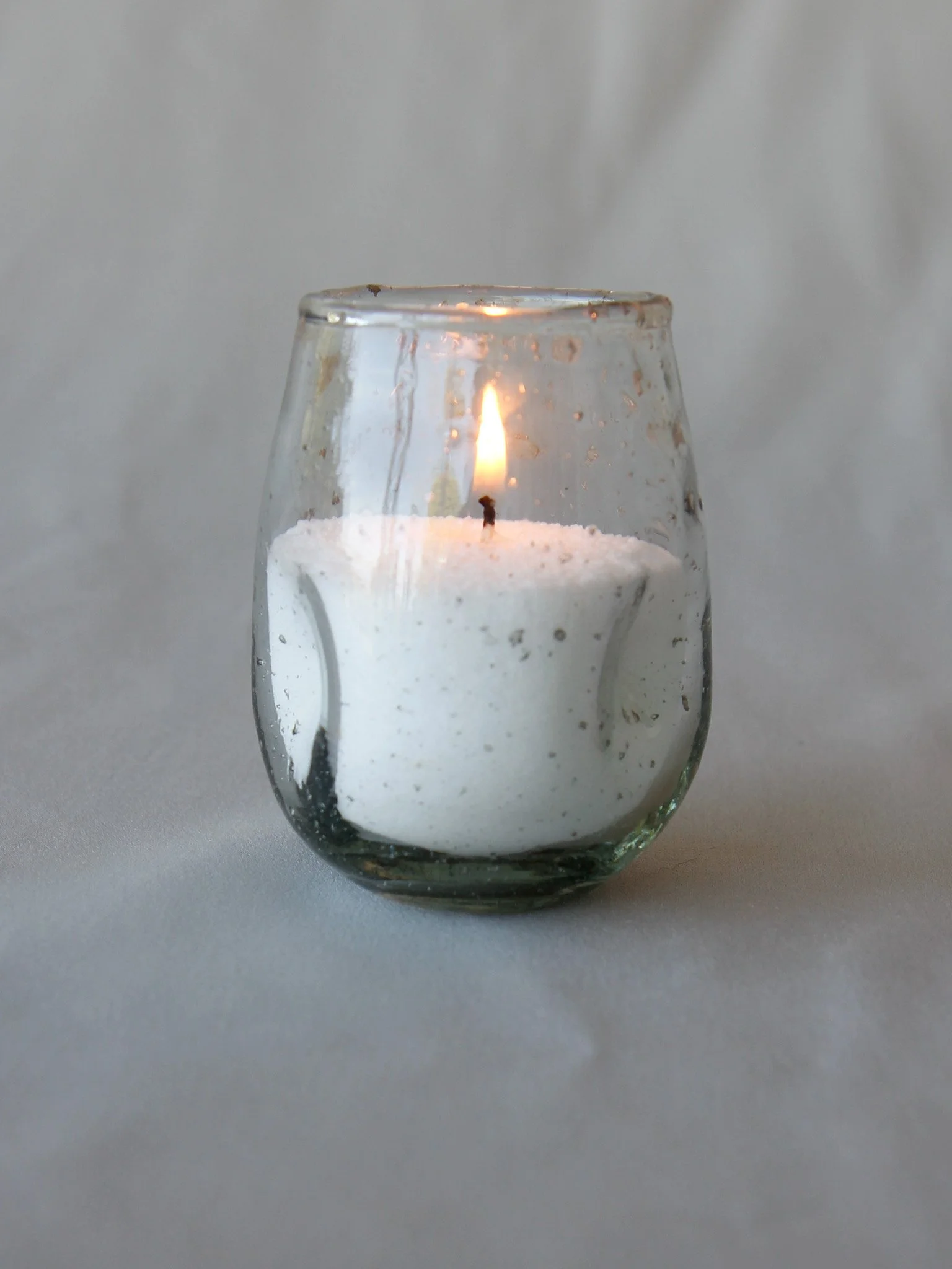 A lit white candle inside a transparent glass holder with water droplets on the outside.