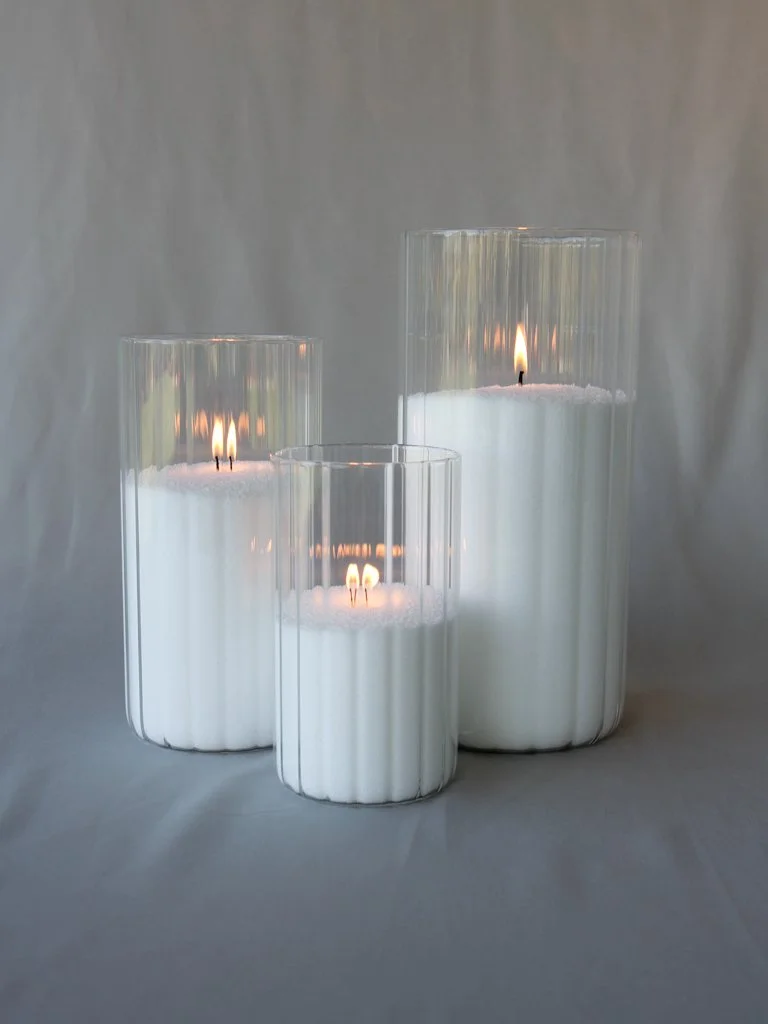 Three tall glass candle holders with white candles inside, lit and burning, placed on a gray surface with a light gray background.