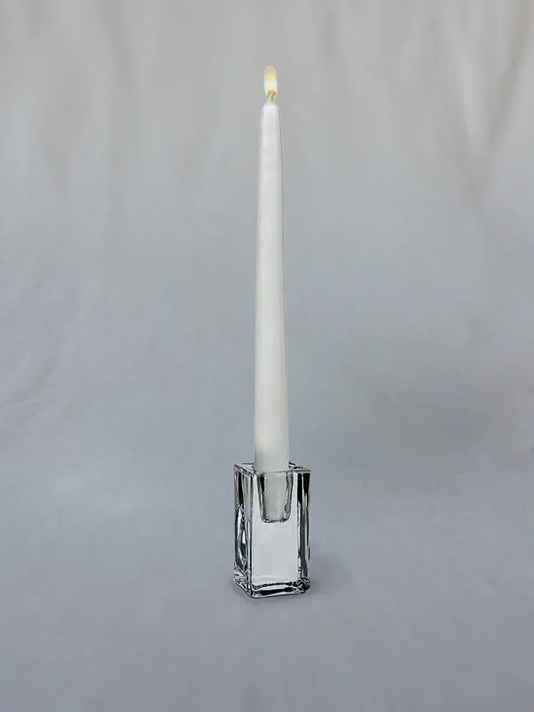 A white candle with a lit wick in a clear glass holder on a plain surface.