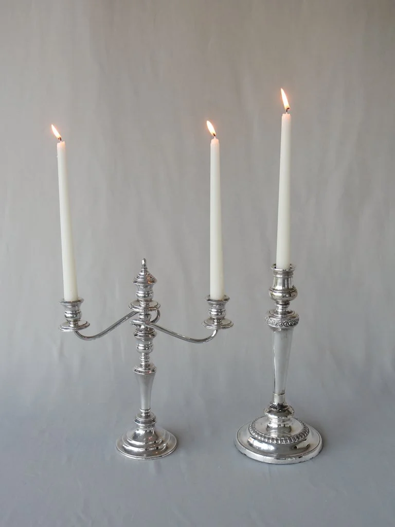 Two silver candelabras with three lit white taper candles on a plain surface with a neutral gray background.