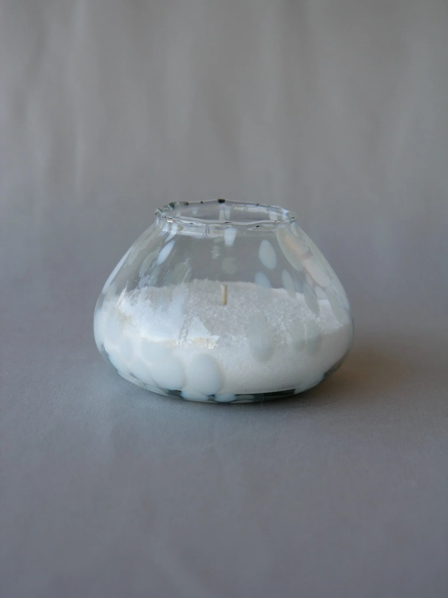 A small round glass candle holder with a white candle inside, covered in a pattern of white and black dots, placed on a gray surface against a plain gray background.