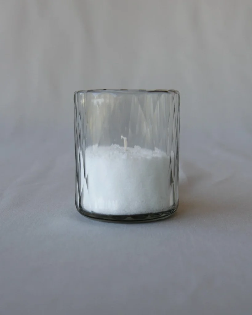 A clear glass container with a textured exterior, filled with white granular substance, likely salt, with an unburned wick inside.