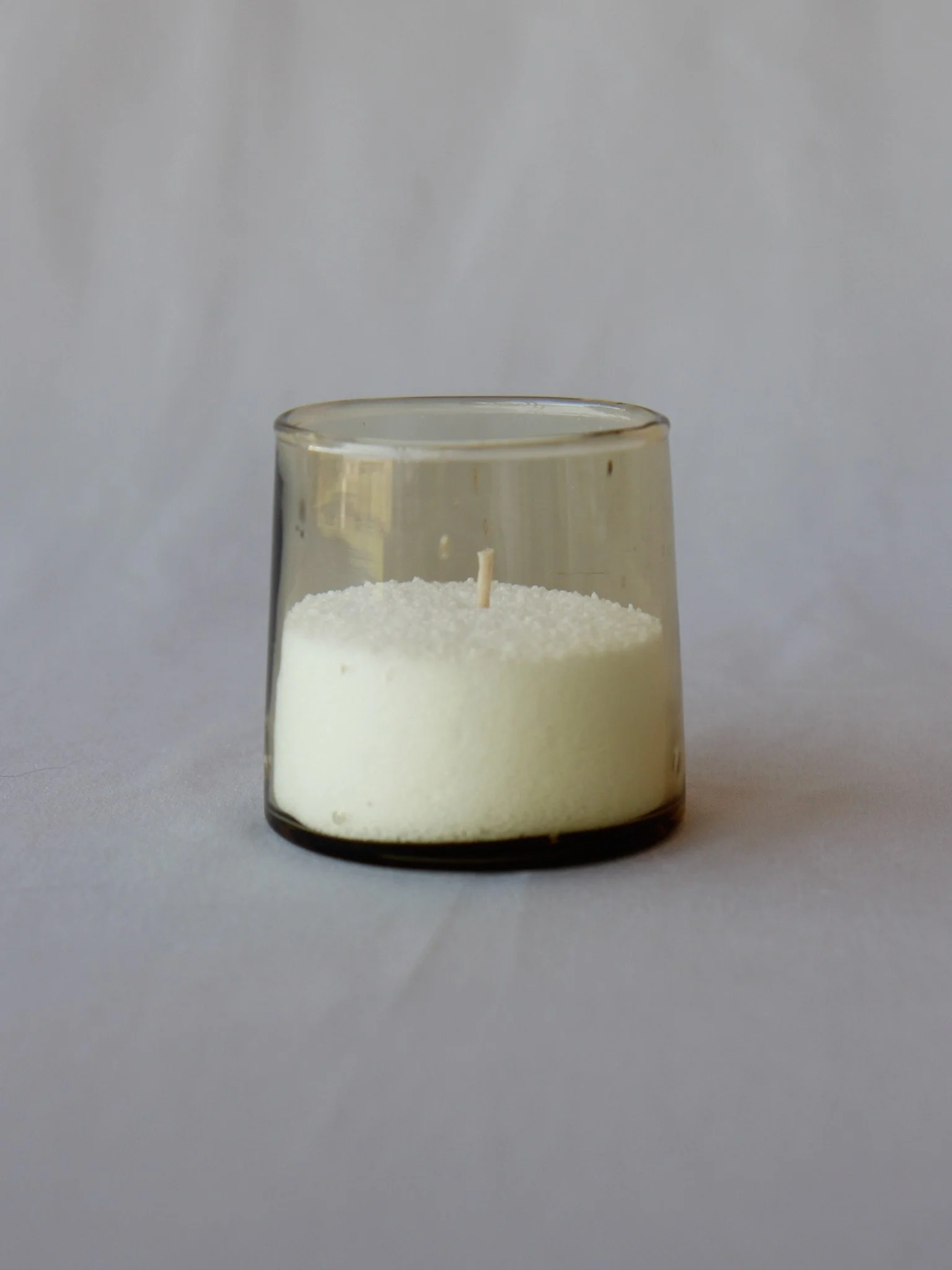 A white candle with a wick inside a transparent glass holder on a light gray surface.