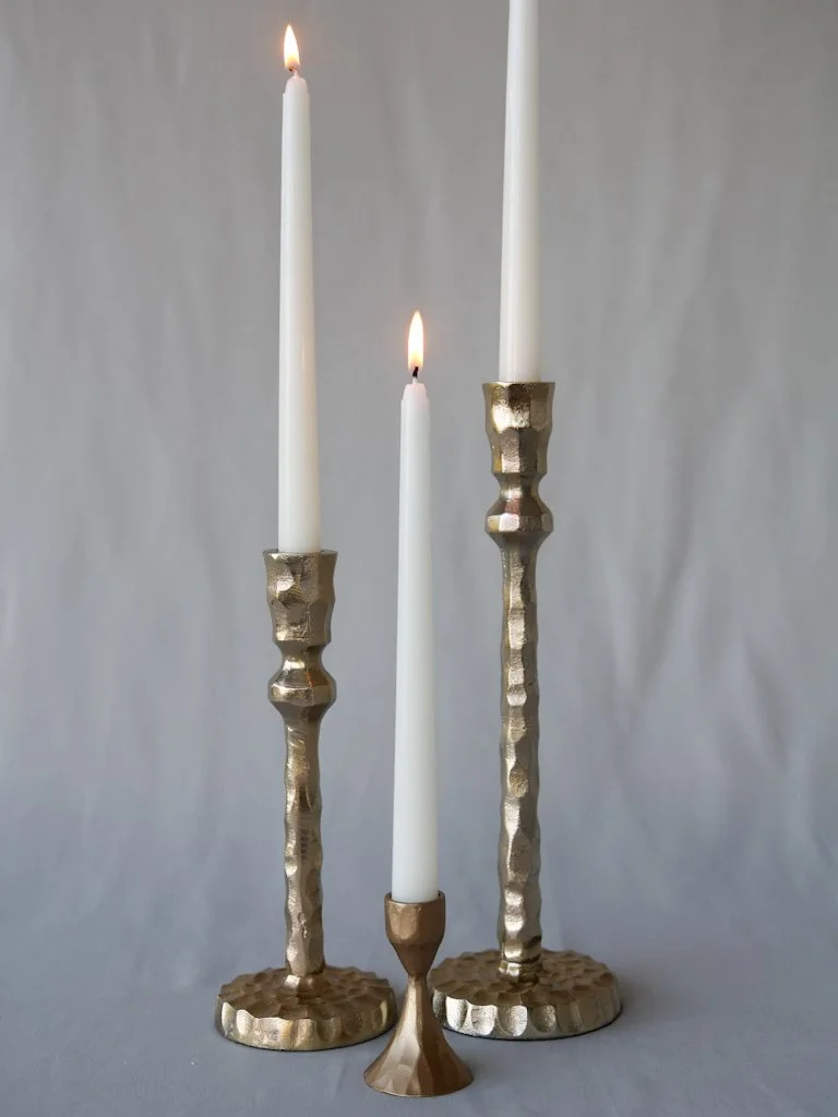 Three tall white taper candles in textured gold candle holders, with two of the candles lit and one unlit, against a plain gray background.