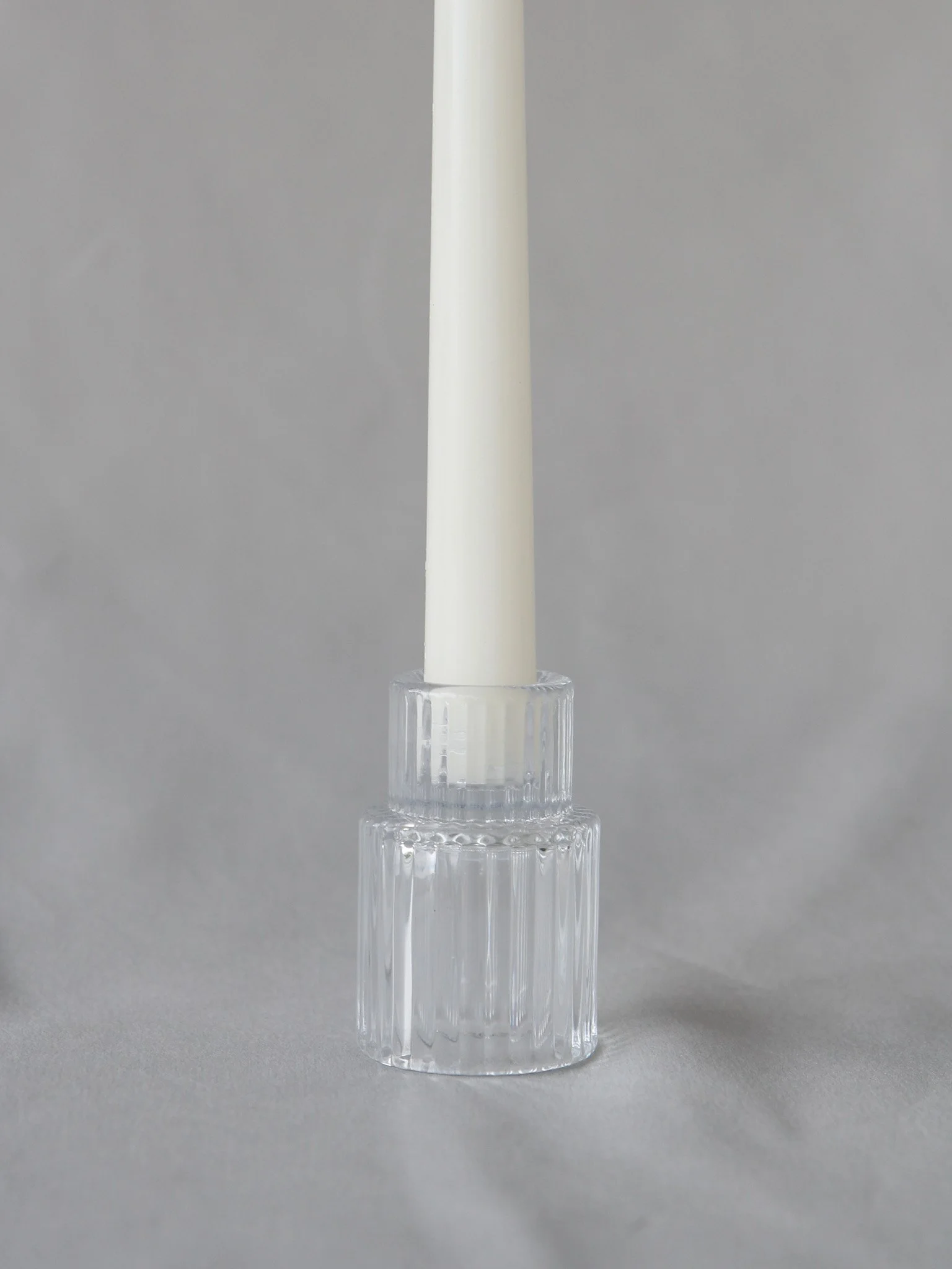 A white taper candle in a clear, ribbed glass candlestick holder against a neutral background.