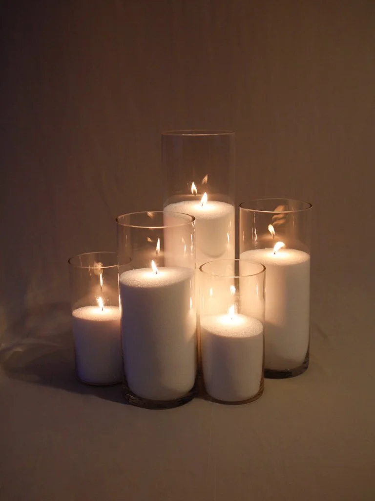 Six white candles in clear glass holders, lit and arranged on a flat surface, casting a soft glow against a dark background.