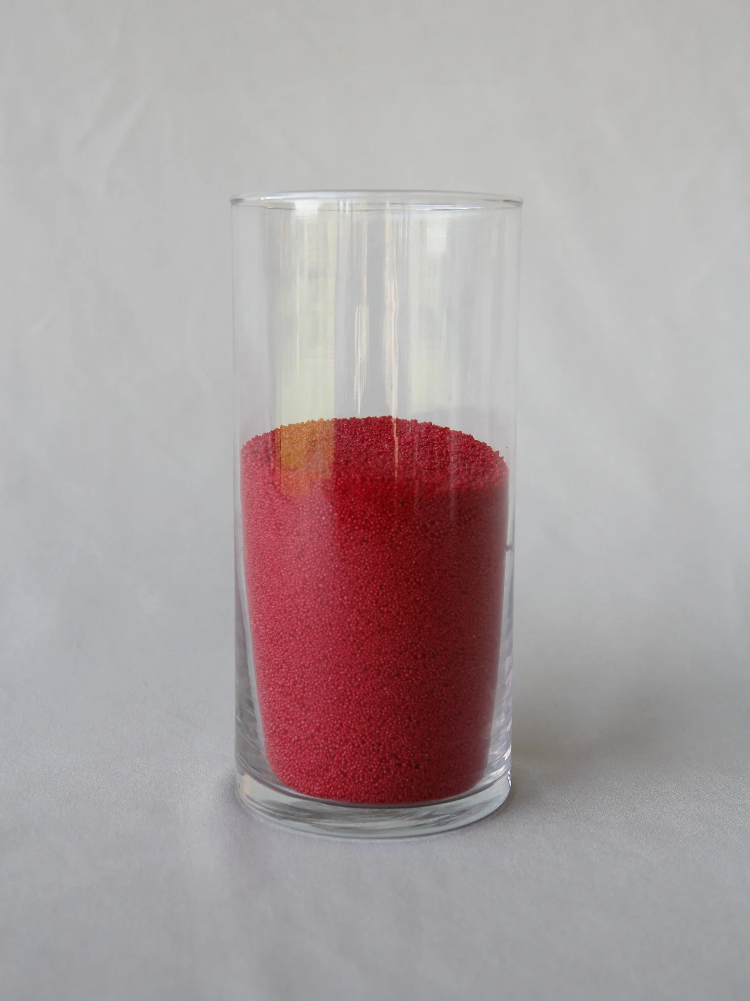 A tall clear glass filled with red and yellow tiny spheres, possibly beads or granules, on a plain white background.