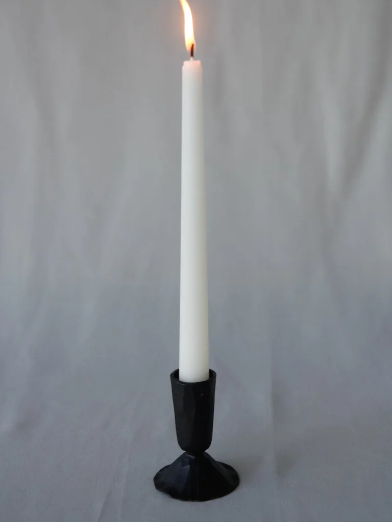 A tall white candle in a black holder with a small flame burning at the top.