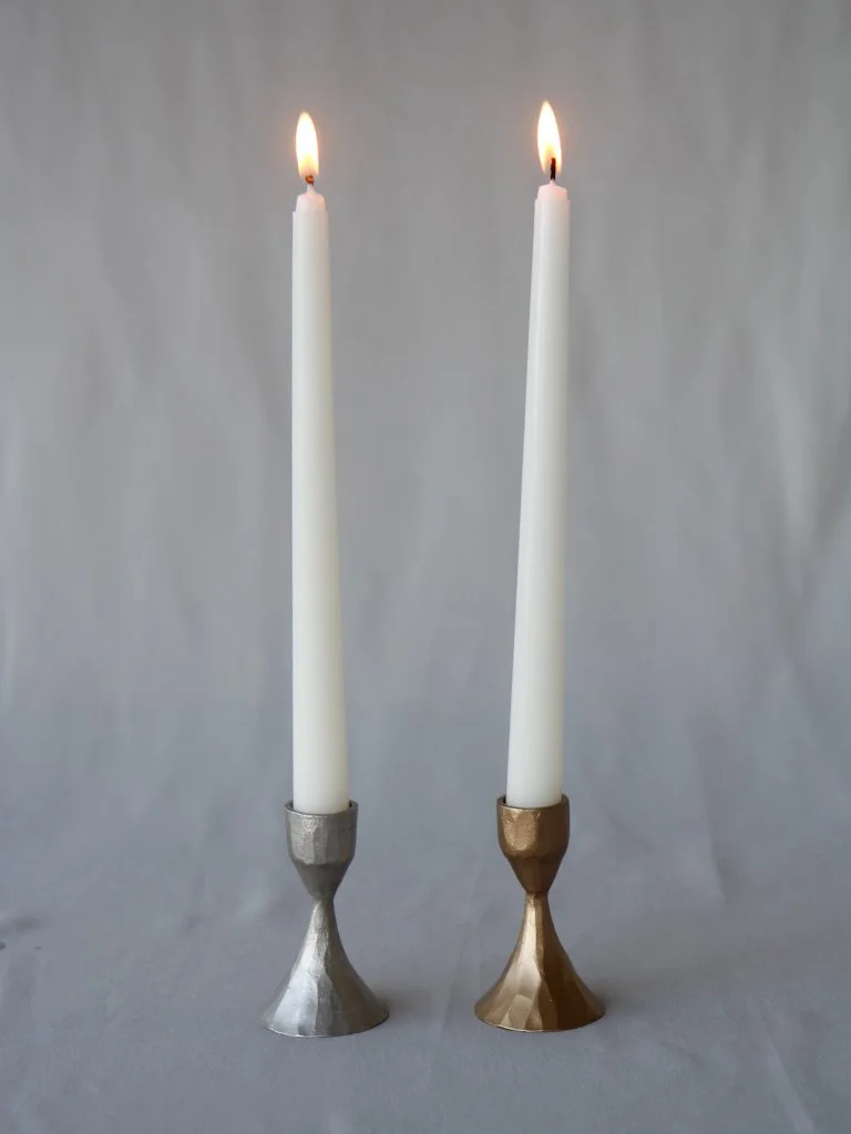 Two taper candles, one silver and one gold, lit and in matching holders, against a plain background.