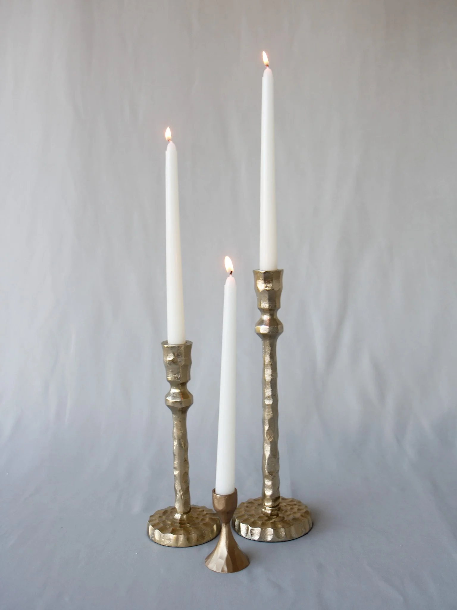 Three lit white candles of varying heights in hammered gold candleholders on a gray background.