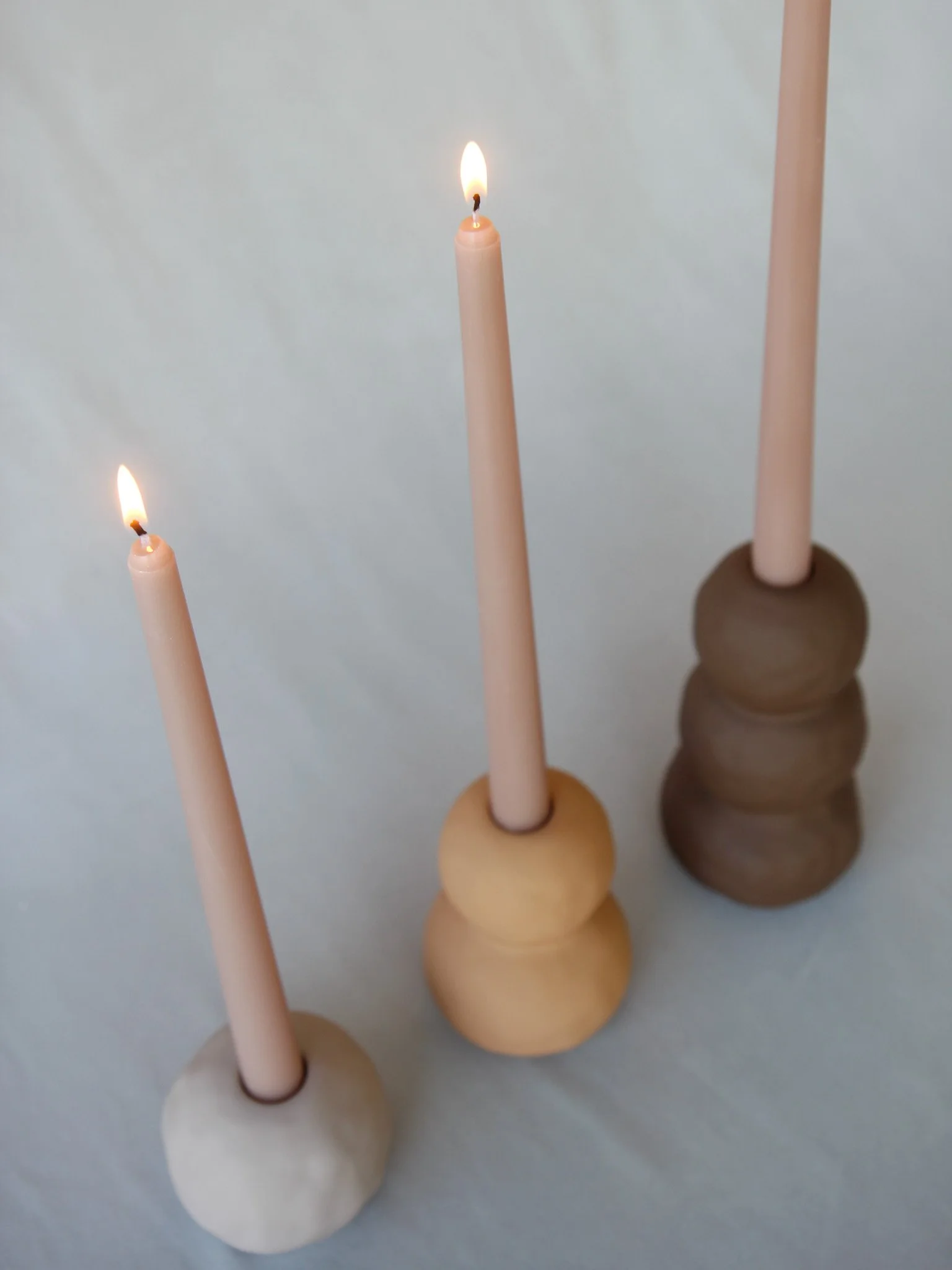 Three beige, brown, and white ceramic candle holders with pink taper candles, two of which are lit, arranged in a row on a light surface.