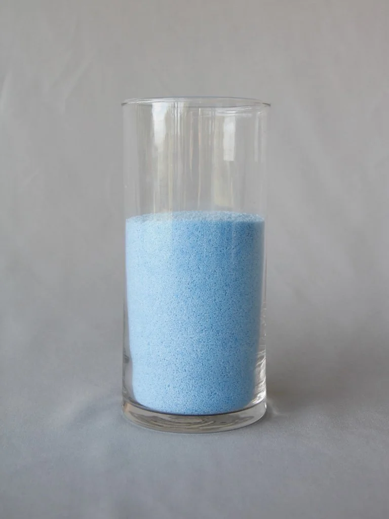 Tall clear glass filled with blue granular substance, set against a plain light gray background.