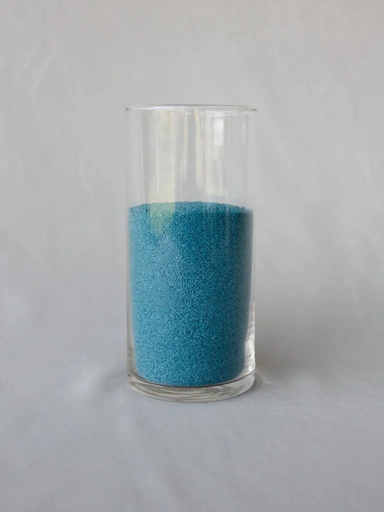 A clear glass filled with small blue beads or granular material against a plain light background.