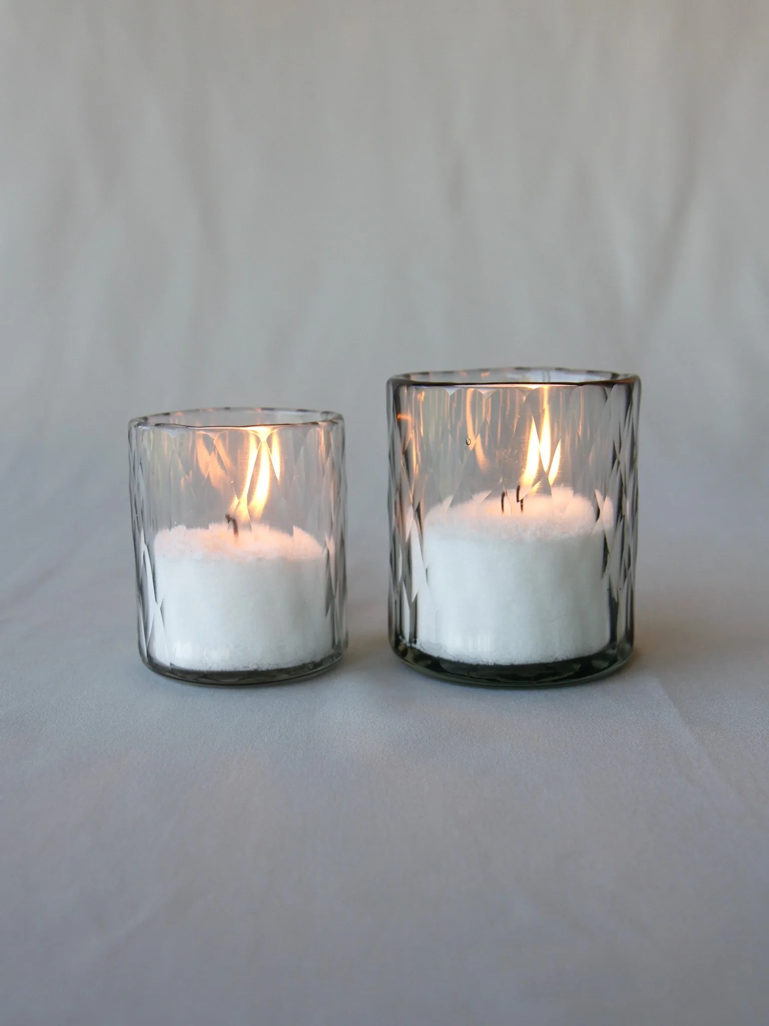 Two lit candles in clear glass holders, one smaller on the left and one larger on the right, placed on a white surface with a plain white background.