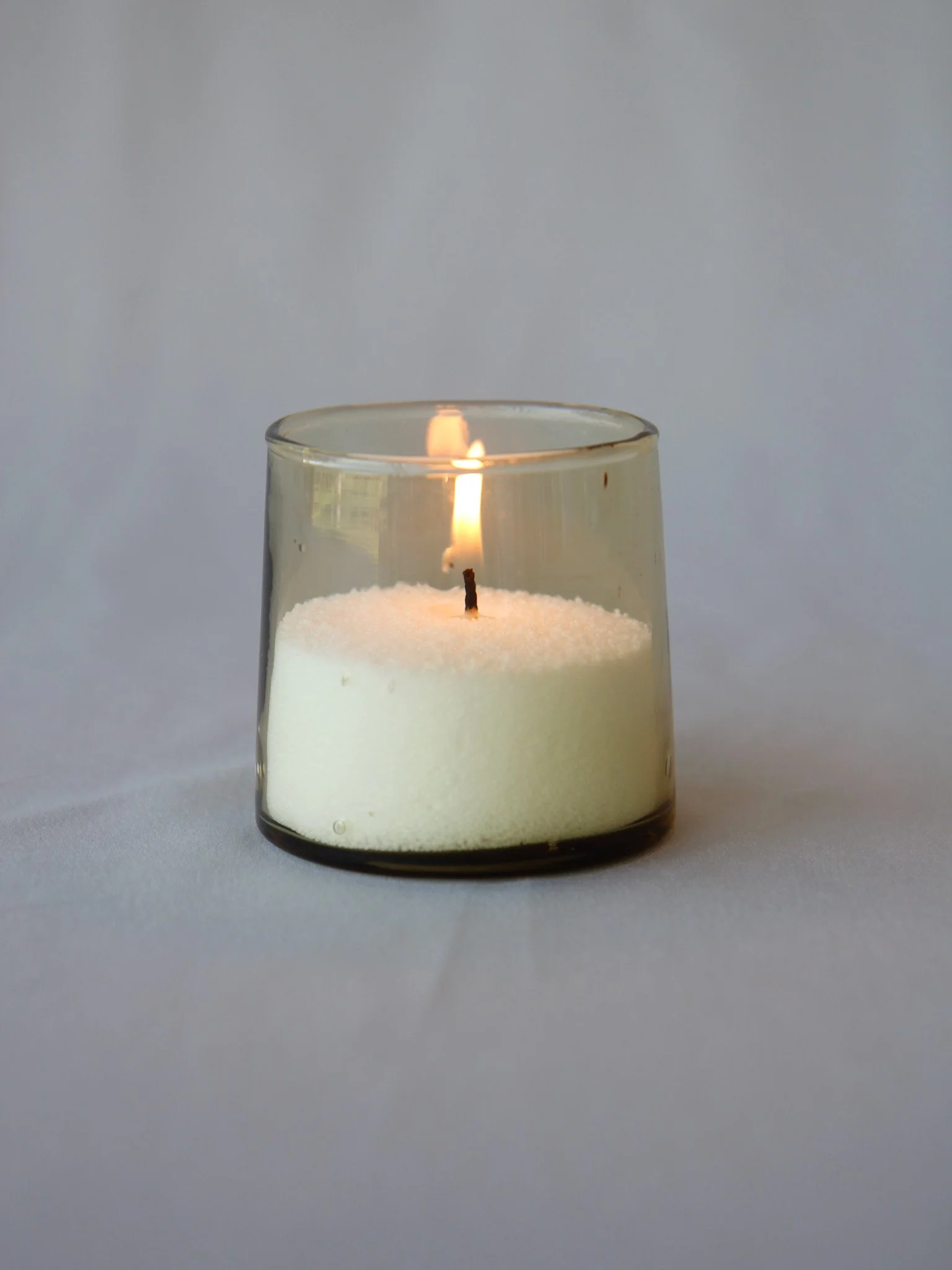 A lit candle in a clear glass holder with a white wax interior on a plain white surface.