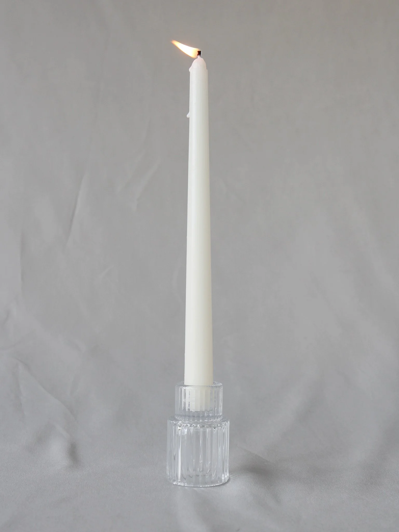A white candle burning in a clear glass holder on a gray surface with a plain gray background.