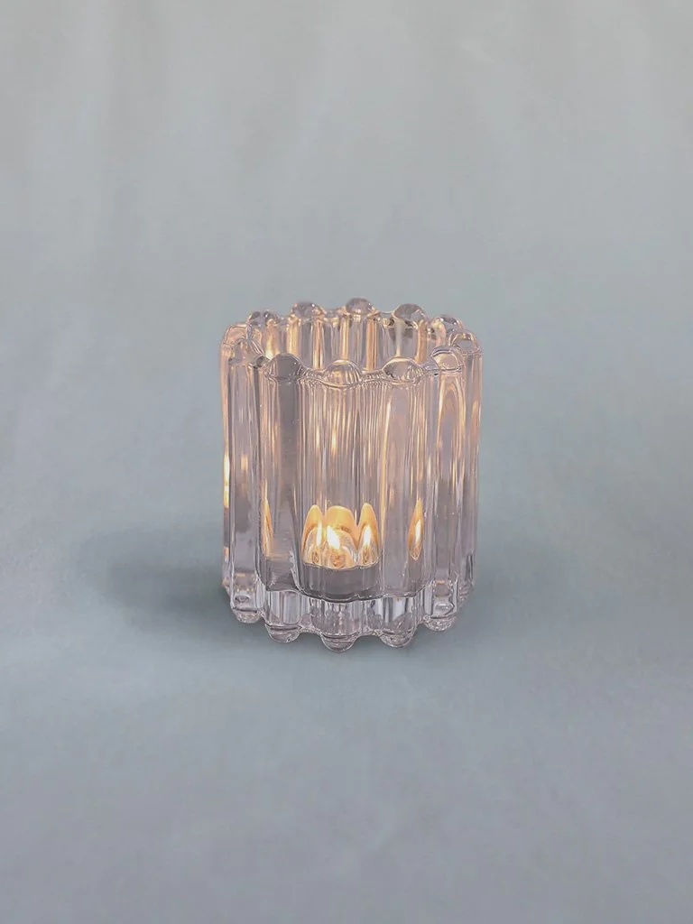 A decorative glass candle holder with a lit candle inside against a plain light background.