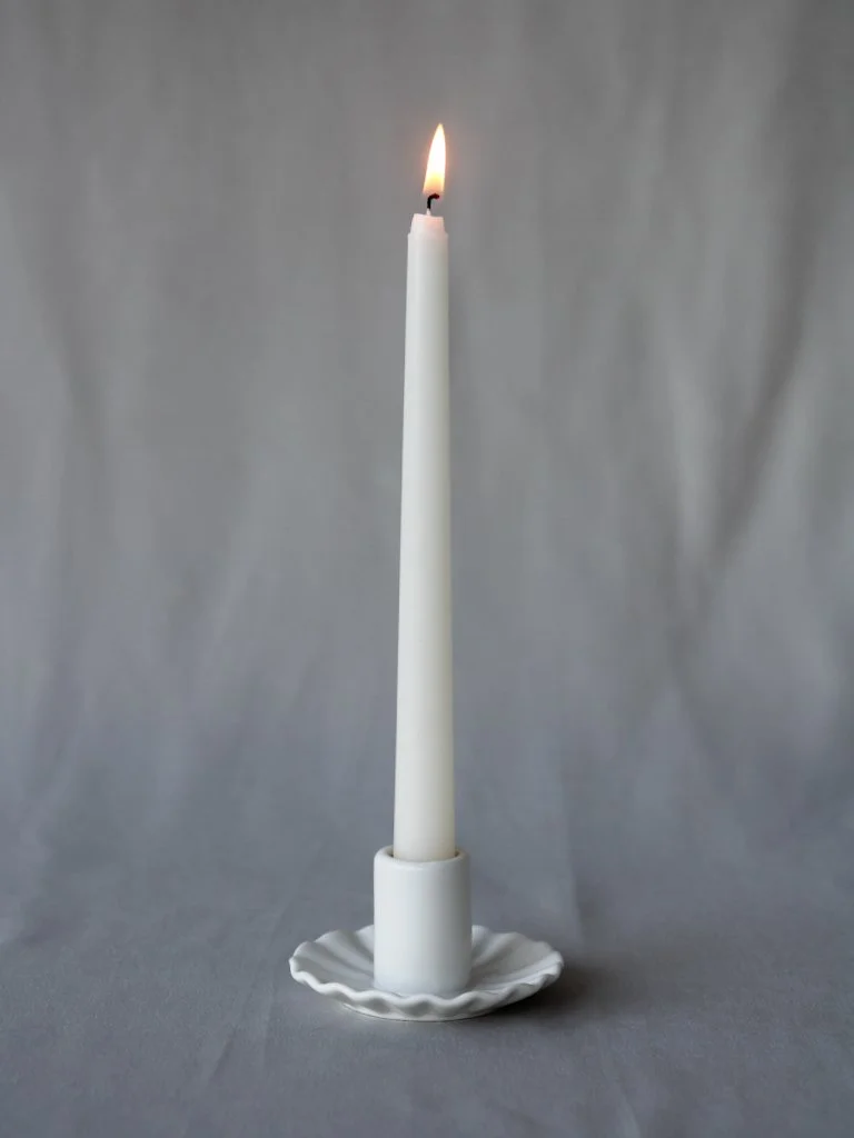 White candle with a small flame on a white, fluted ceramic holder against a gray background.