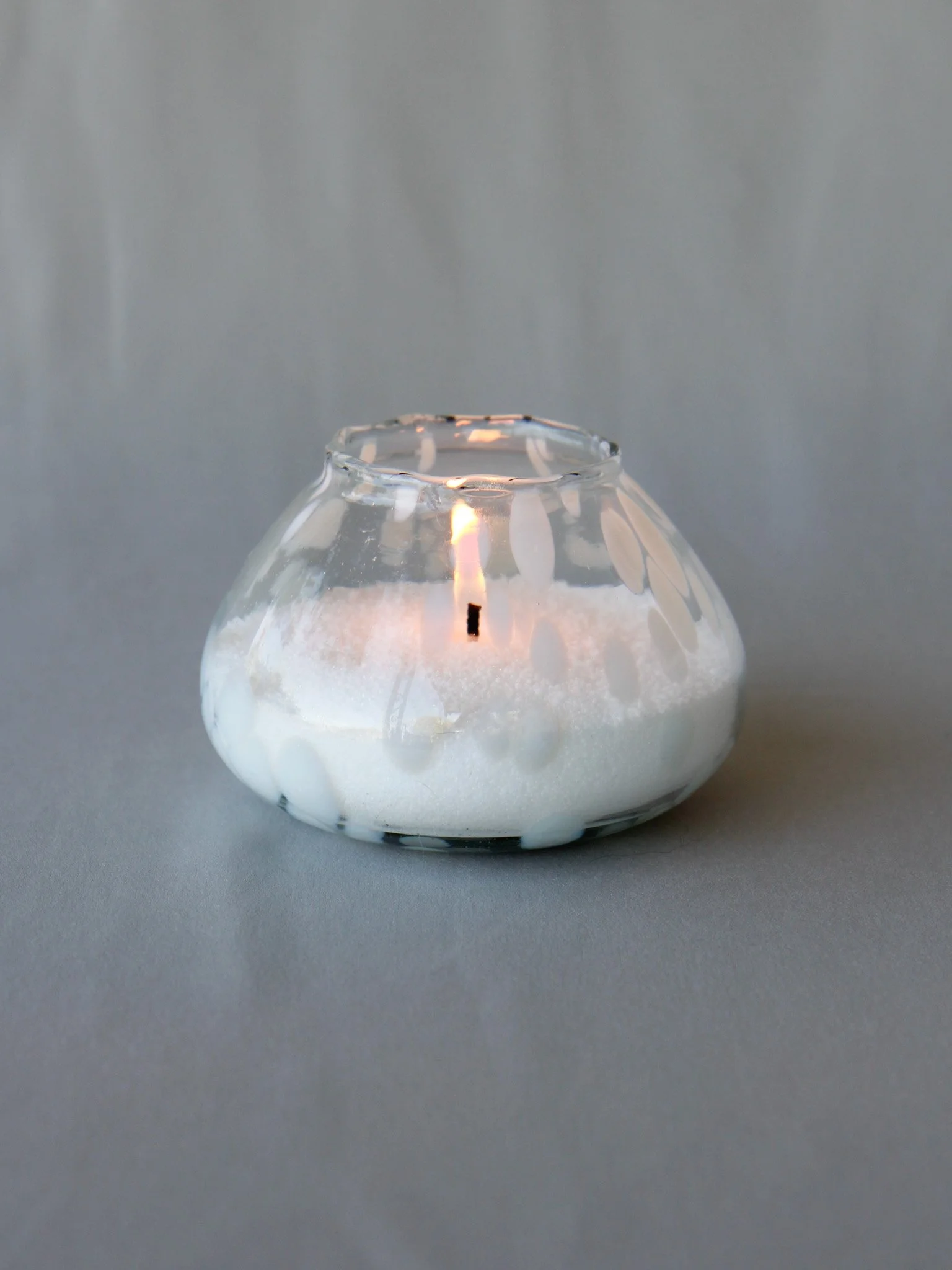 A small glass candle holder with a burning candle inside, surrounded by white decorative salt or sand.