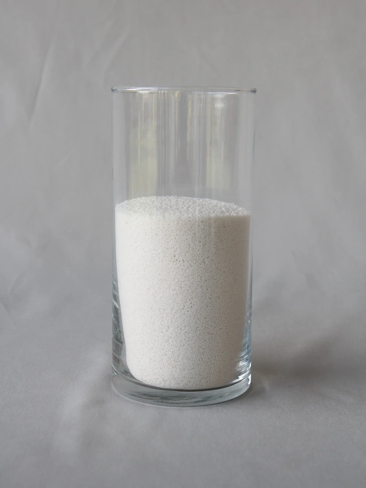 A tall, clear glass container filled with white granular powder, set against a plain gray background.