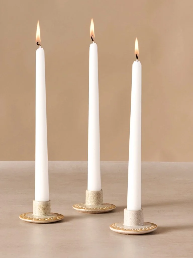 Three white taper candles with flames, placed in gold-colored candle holders on a light-colored surface.