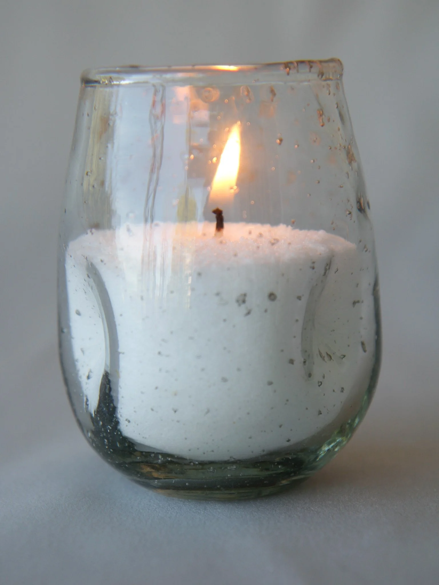 Close-up of a glass candle holder with a lit white candle inside, with a visible flame and some melted wax.