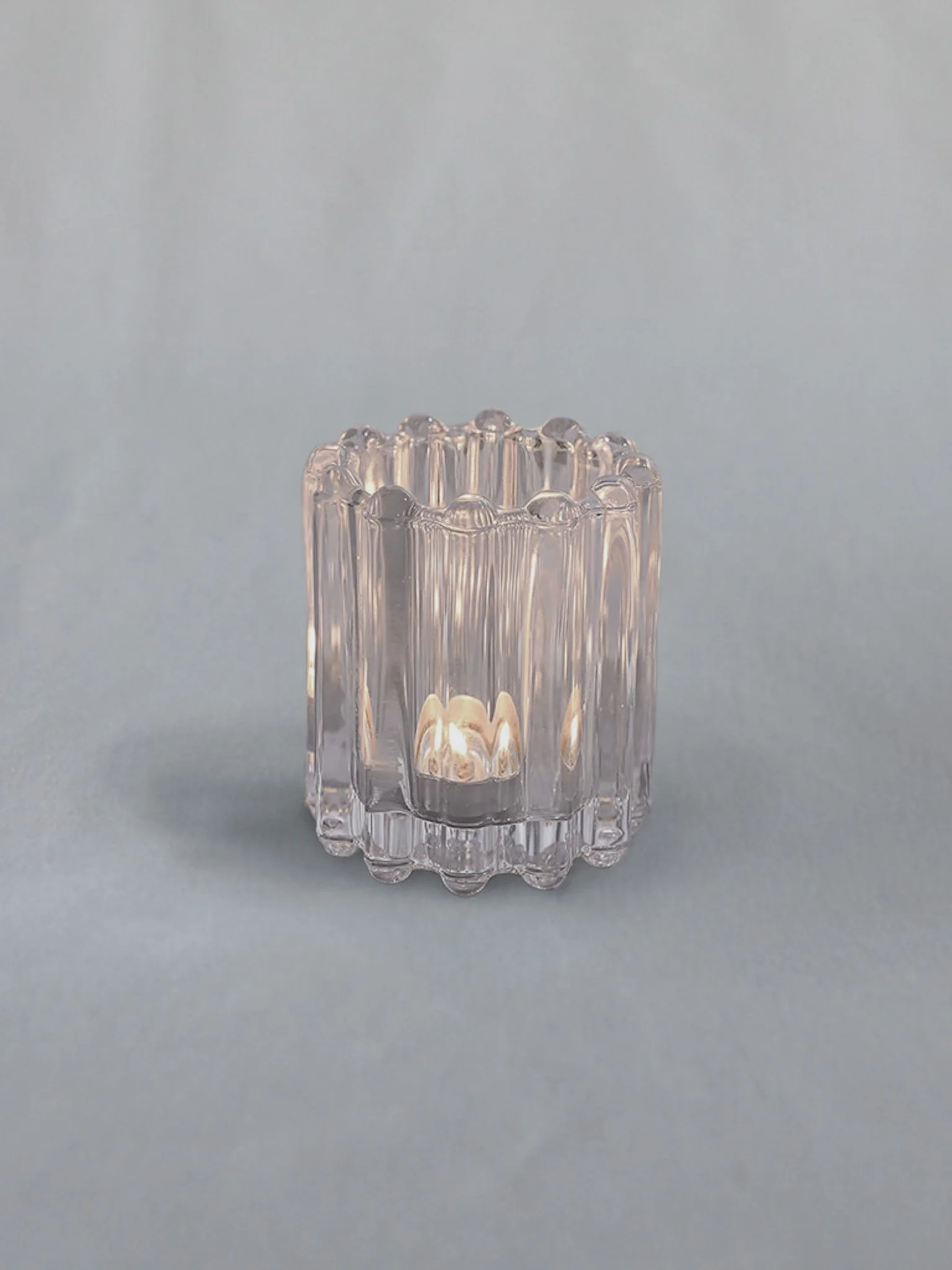 A small, clear, ridged glass candle holder with a lit candle inside, placed on a plain, light-colored background.