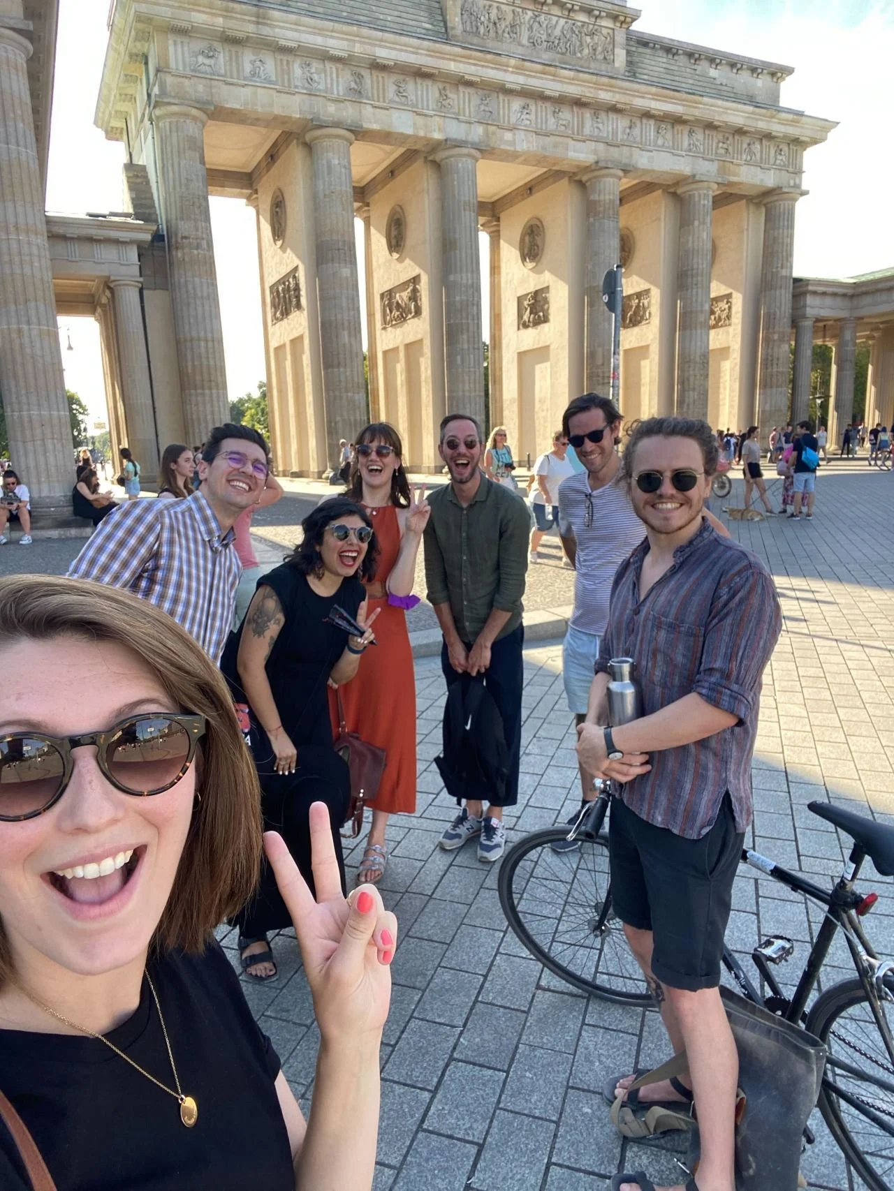 A group of seven friends taking a selfie in front of the Brandenburg Gate in Berlin, Germany, on a sunny day. They are smiling and making peace signs, with the historic monument and other visitors in the background.