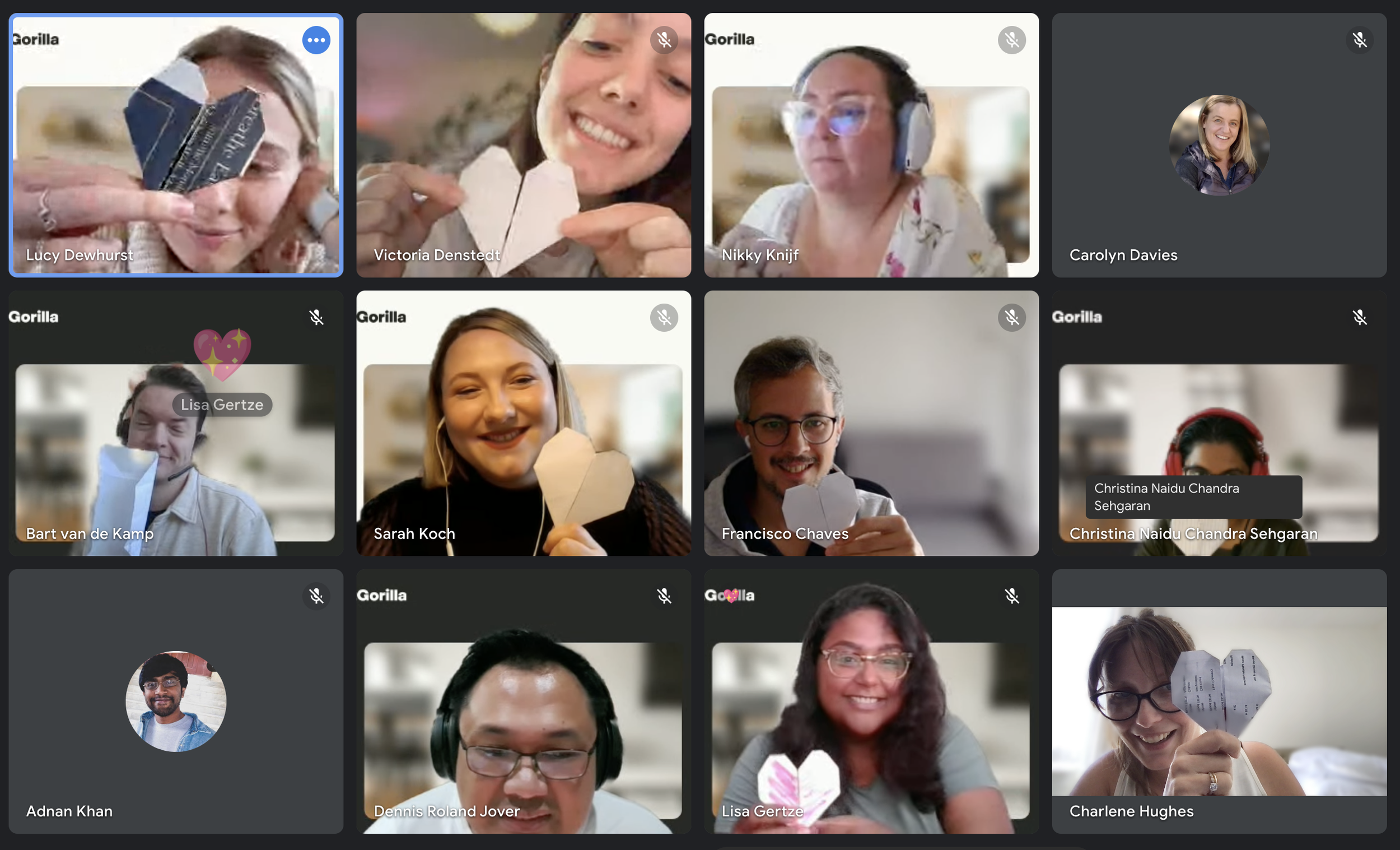 A virtual video call with multiple participants, each holding a paper heart, smiling or engaged, with some wearing headphones or glasses.