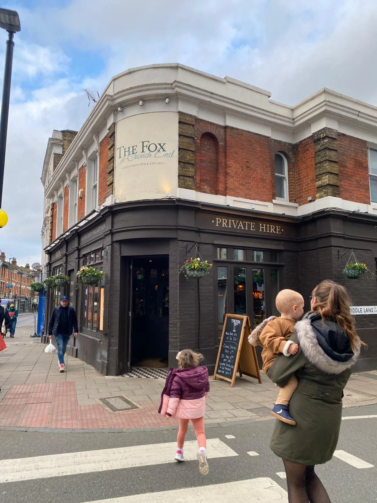 Street corner view of The Fox pub with black exterior, hanging flower baskets, and people walking, including a woman holding a child and a girl crossing the crosswalk.