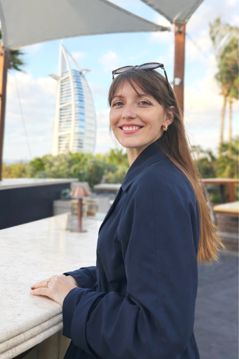A woman with long brown hair, wearing a dark blazer and sunglasses on her head, smiling outdoors at a rooftop venue with the Burj Al Arab in Dubai in the background.