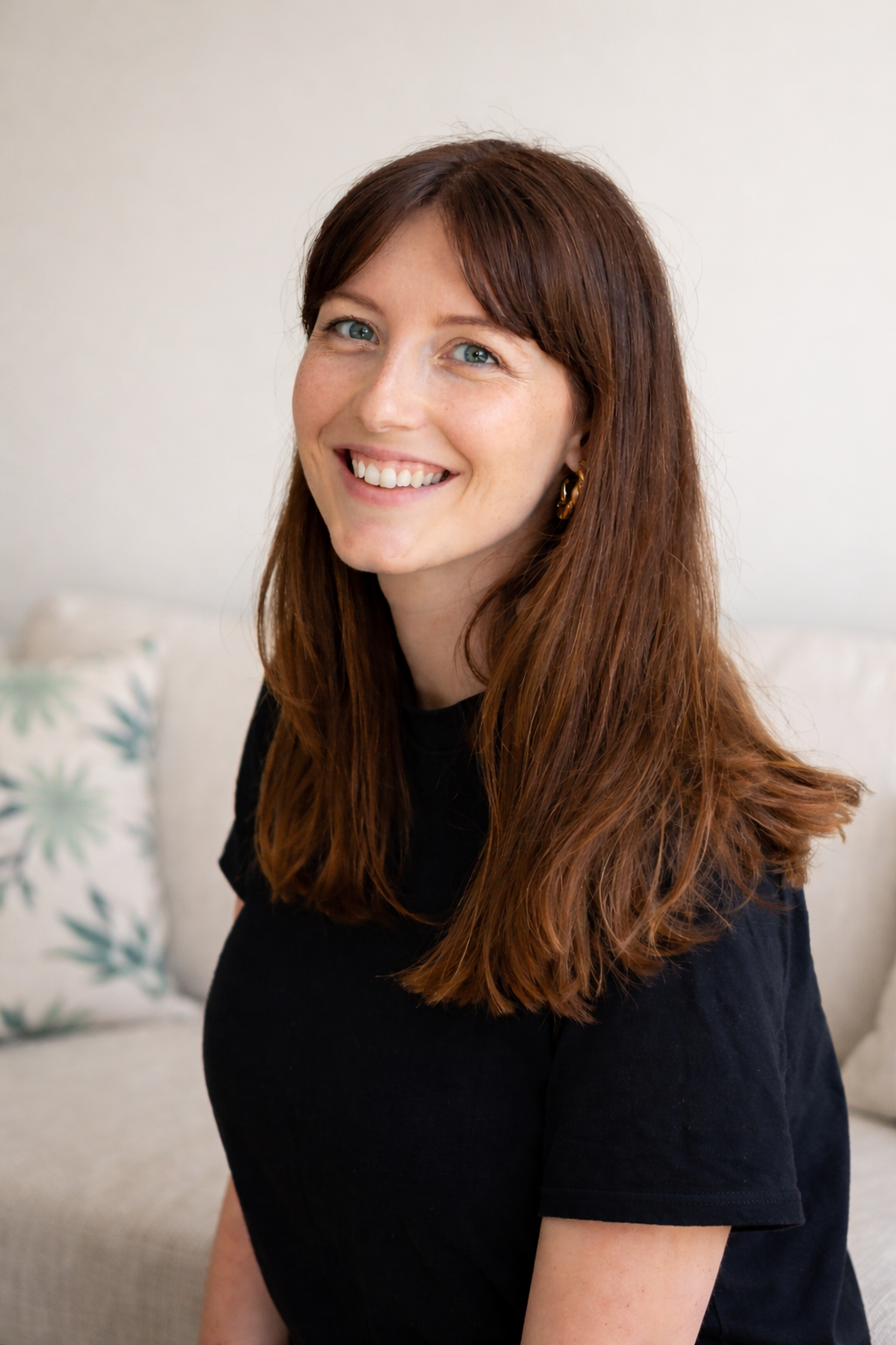 A woman with long reddish-brown hair, blue eyes, and earrings smiling at the camera, sitting on a beige couch with patterned pillows in the background.