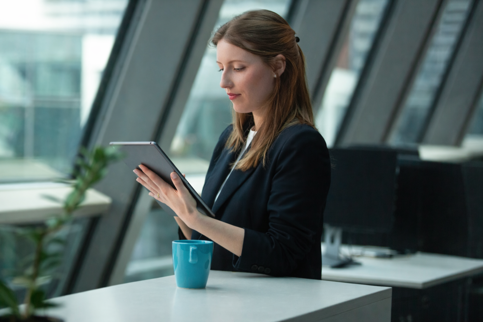 A woman in business attire standing at a white table, looking at her tablet, with a blue coffee mug in front of her, in a modern office with large windows.