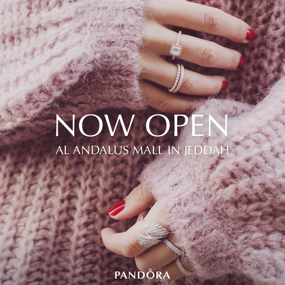 Close-up of a woman's hand showing multiple rings, with long red painted nails, wearing a pink fuzzy sweater. Text overlay states 'NOW OPEN' at AL ANDALUS MALL IN JEDDAH and includes the Pandora logo.