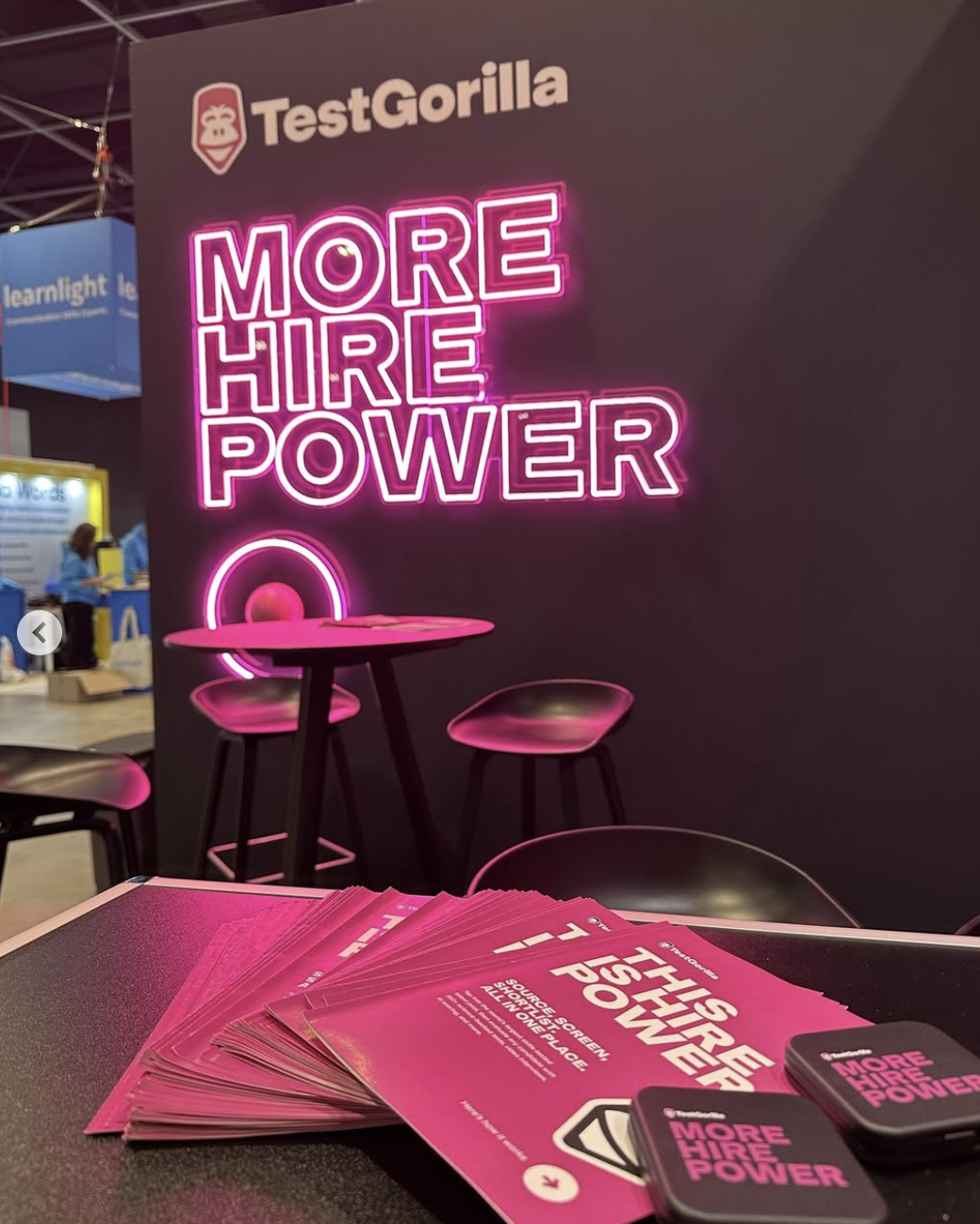 Bright pink neon sign displaying 'More Hire Power' at TestGorilla booth with pink flyers on a table and black chairs around.