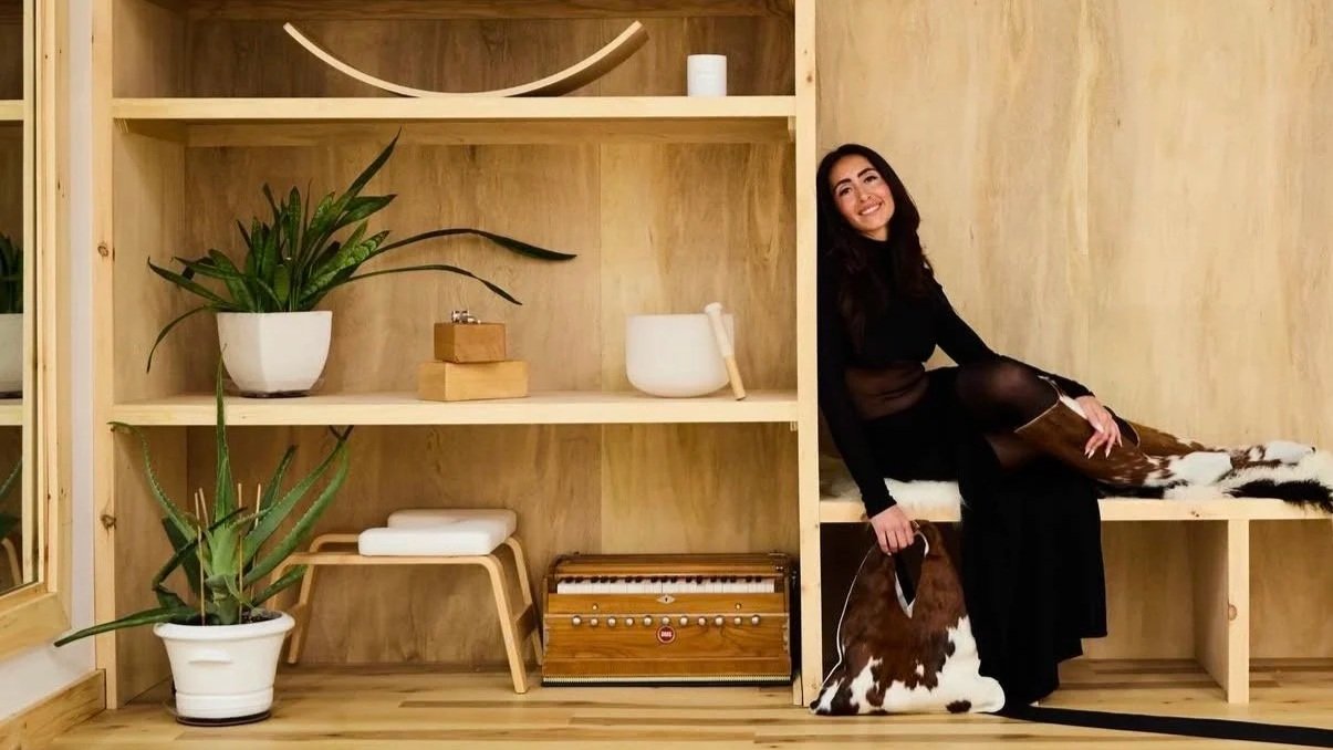Lauren B Yoga, young woman with long dark hair smiling and sitting on a wooden bench covered with a cowhide rug, in her yoga studio. She is wearing a dress and shiny  tights, holding a phone, a pair of glasses. Yoga mats, sound bowls, blocks, plants.